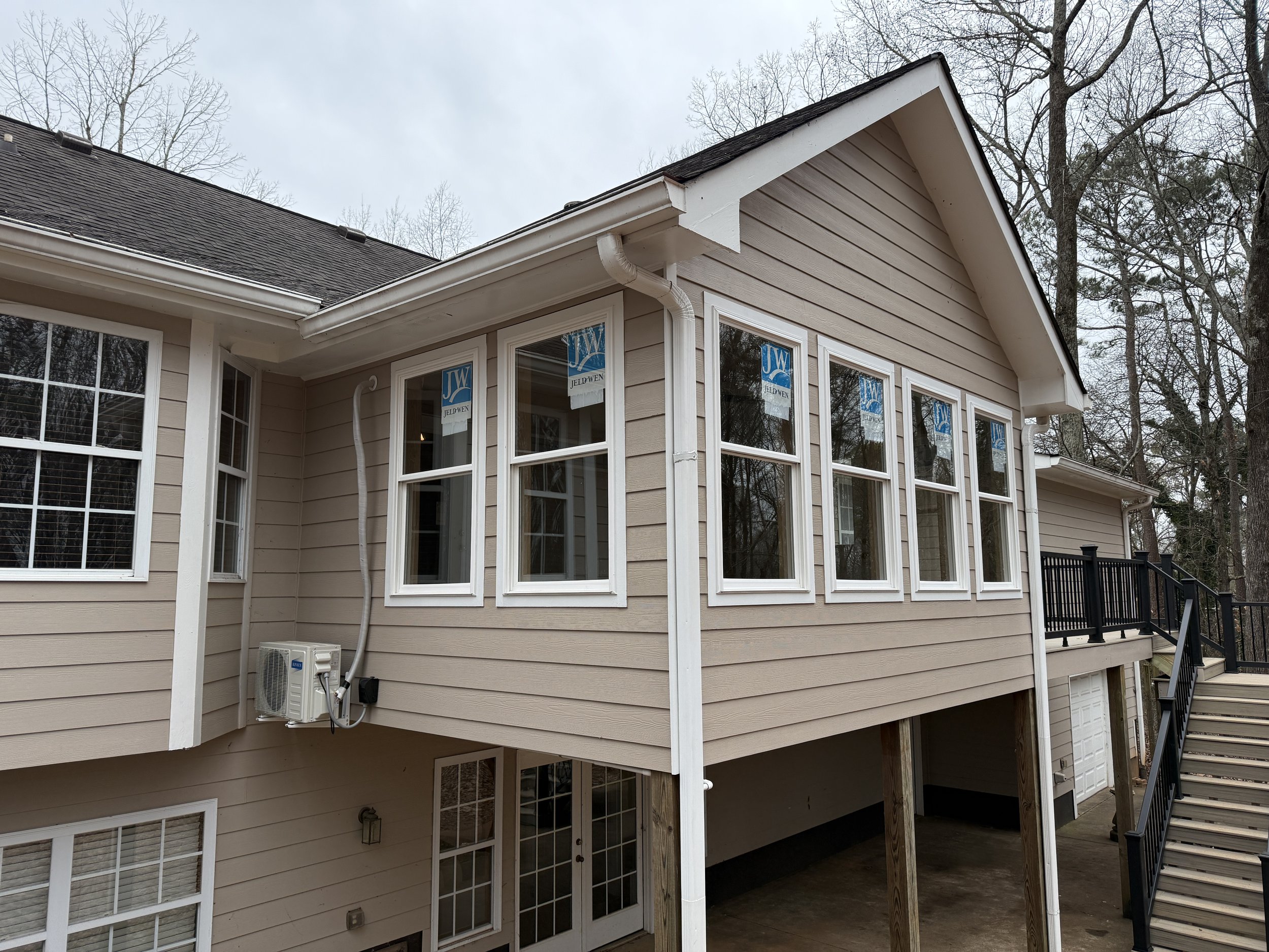 The image shows the exterior of a beige house with multiple white-framed windows. The house has a gabled roof with dark shingles and some trees are visible in the background. There is an air conditioning unit and a gutter system attached to the house, as well as stairs leading up to a door.