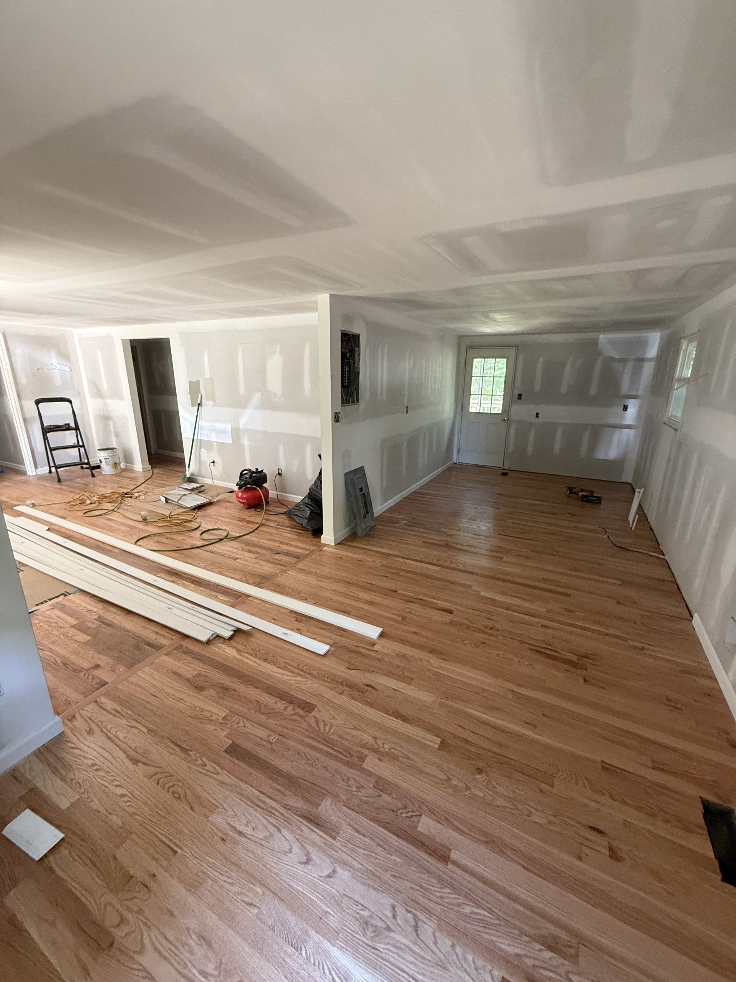 Living room in the process of being renovated, with new hardwood floors installed, unassembled trim pieces on the floor, tools and construction materials scattered around, unfinished walls, and a small window letting in natural light.