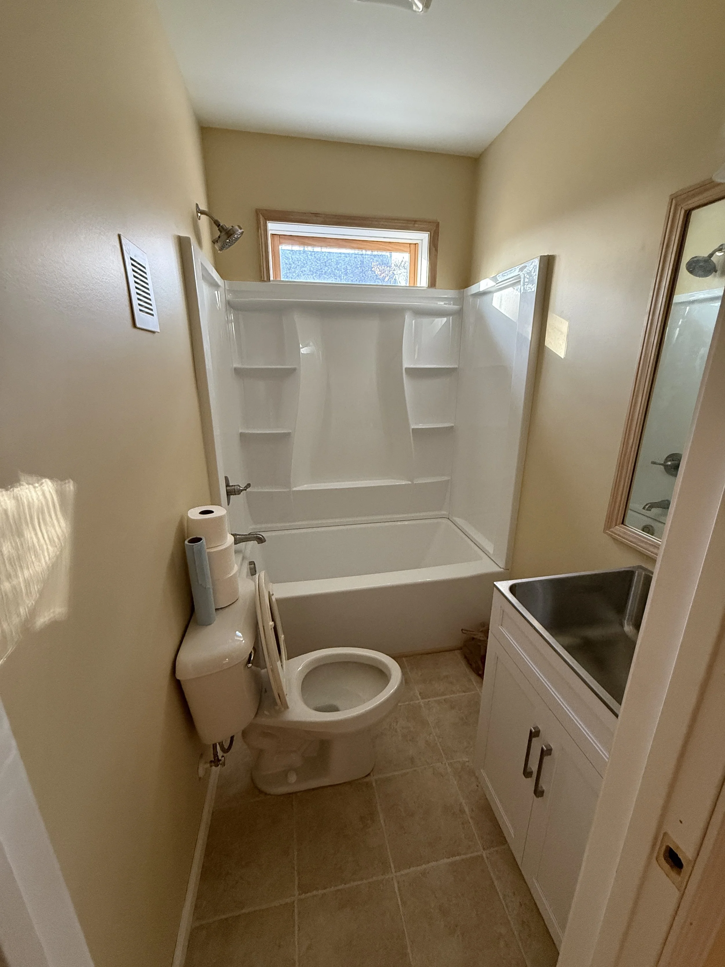 Small bathroom with a white toilet, a white sink with a small cabinet below, a mirror, beige walls, a small window, a shower with shelves, and a toilet paper roll on the side.