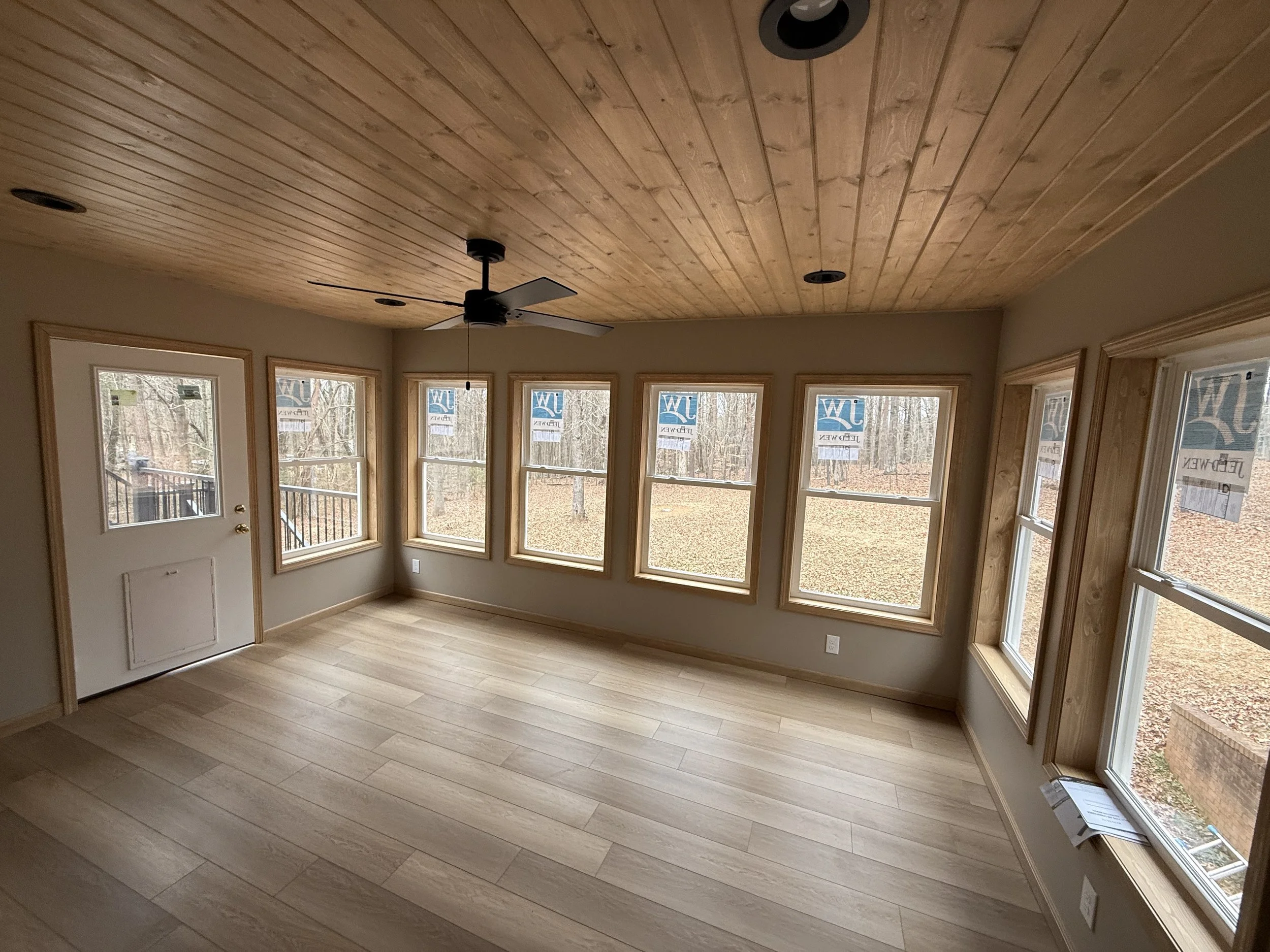 Interior of a room with wooden ceiling, multiple windows, a ceiling fan, and a front door with glass panels.