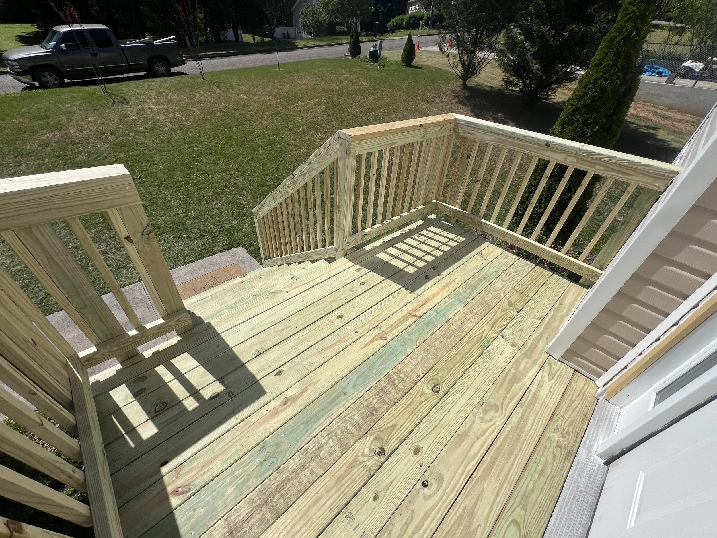 Newly built wooden deck with railing attached to a house, overlooking a green lawn with trees and a driveway with a parked pickup truck.