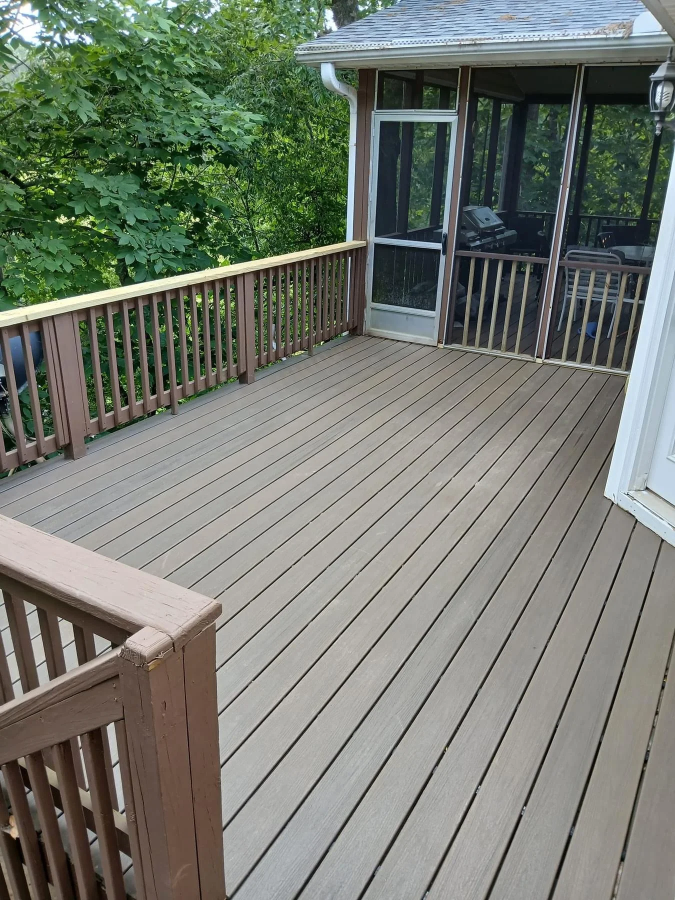 A wooden deck with brown railing, adjacent to a screened porch, with a view of green trees and bushes nearby.