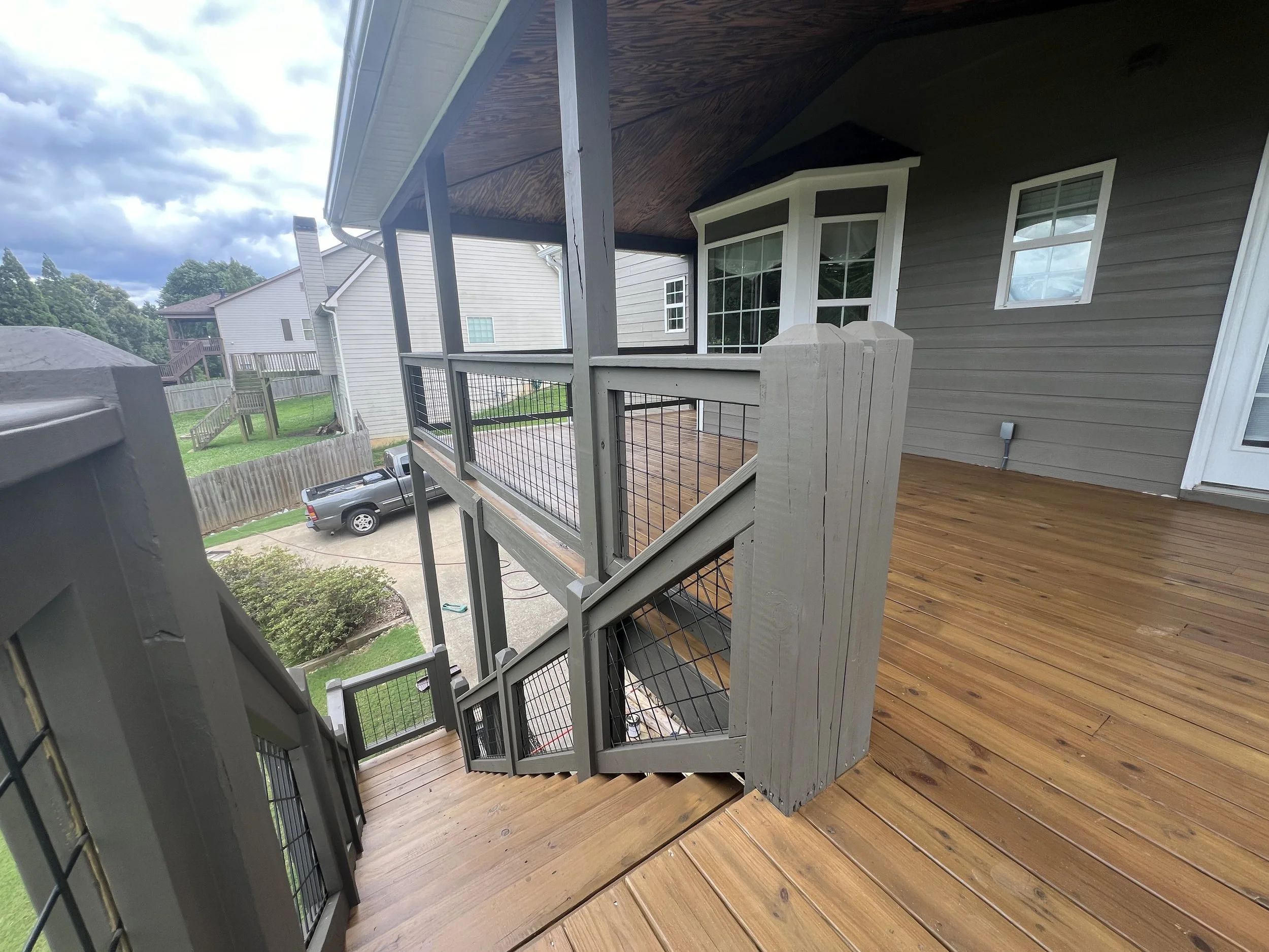 Wooden deck with stairs leading down to a backyard with lawn, bushes, and a driveway. Houses with siding and fences are visible in the background.