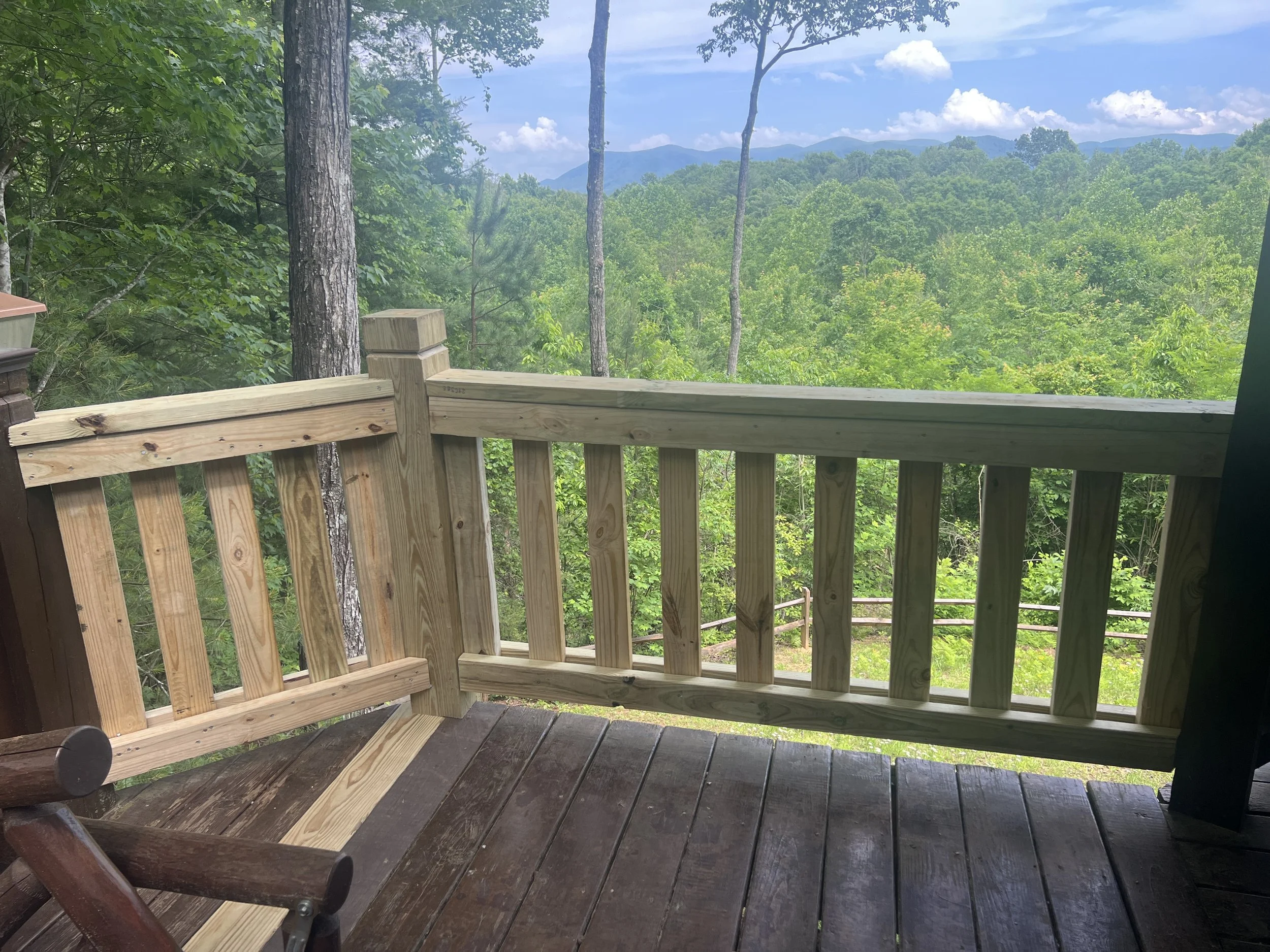 Wooden balcony overlooking a lush green forest with distant mountains and a partly cloudy sky.