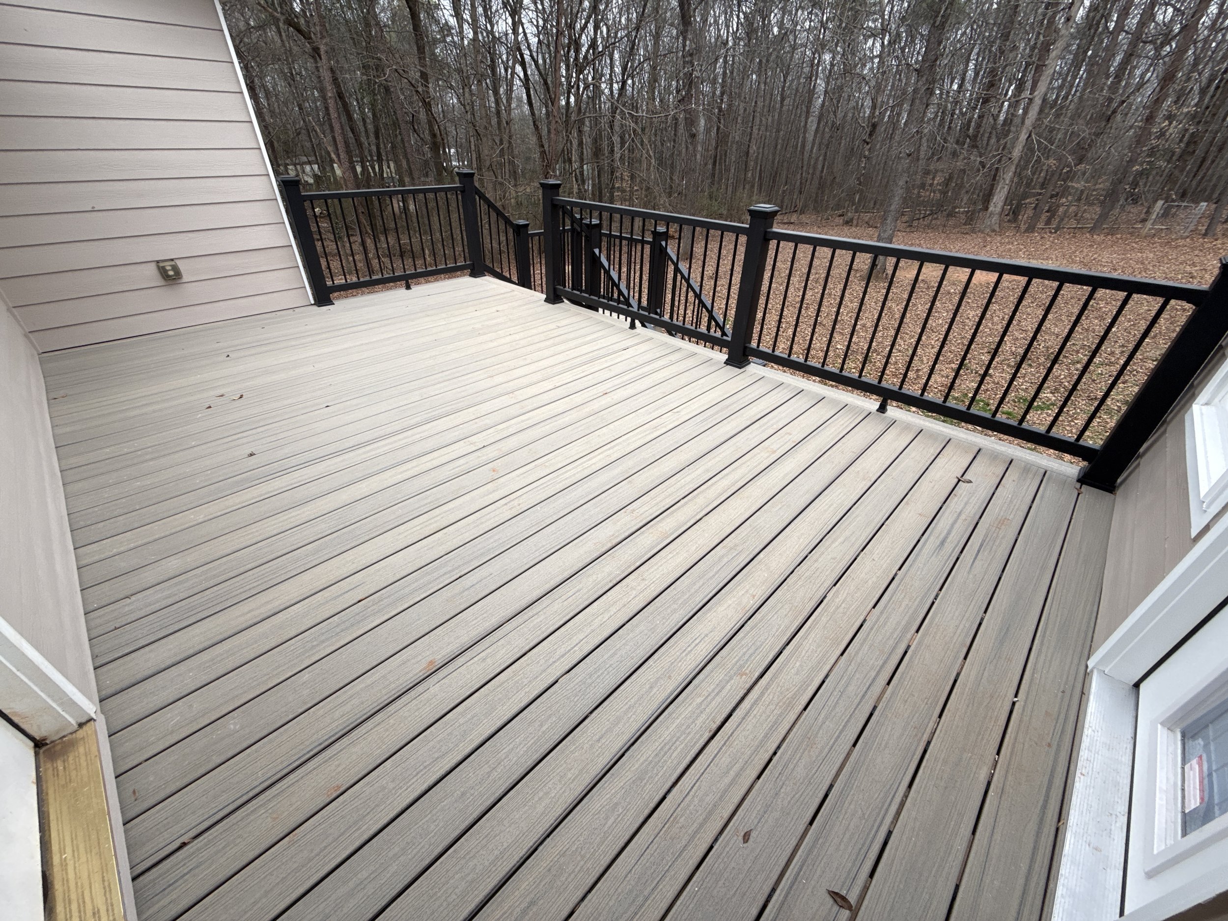 A view of a wooden deck attached to a house, with a black metal railing overlooking a wooded area with leafless trees and brown leaves on the ground.
