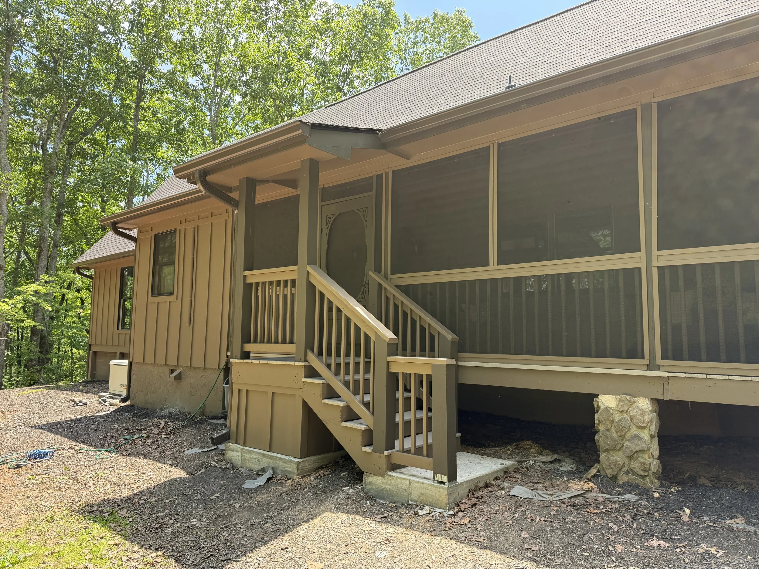 Exterior view of a house with a screened porch, wooden stairs, surrounded by trees, with a gravel yard and stone foundation.