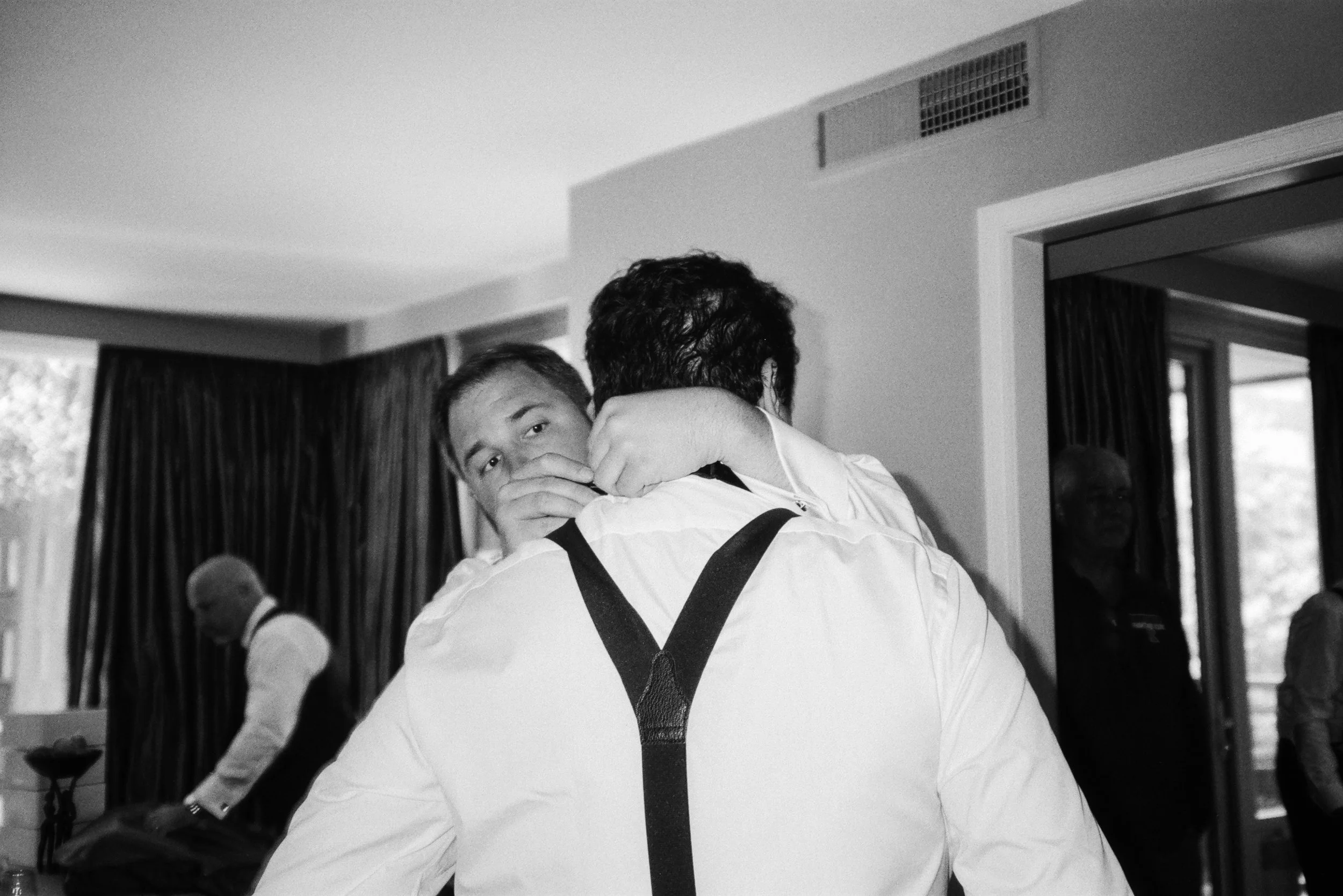 Two men dressed in formal attire, possibly during a special event, embrace each other. The man facing the camera has short hair and a serious expression. The background shows a room with large windows, curtains, and other people present.