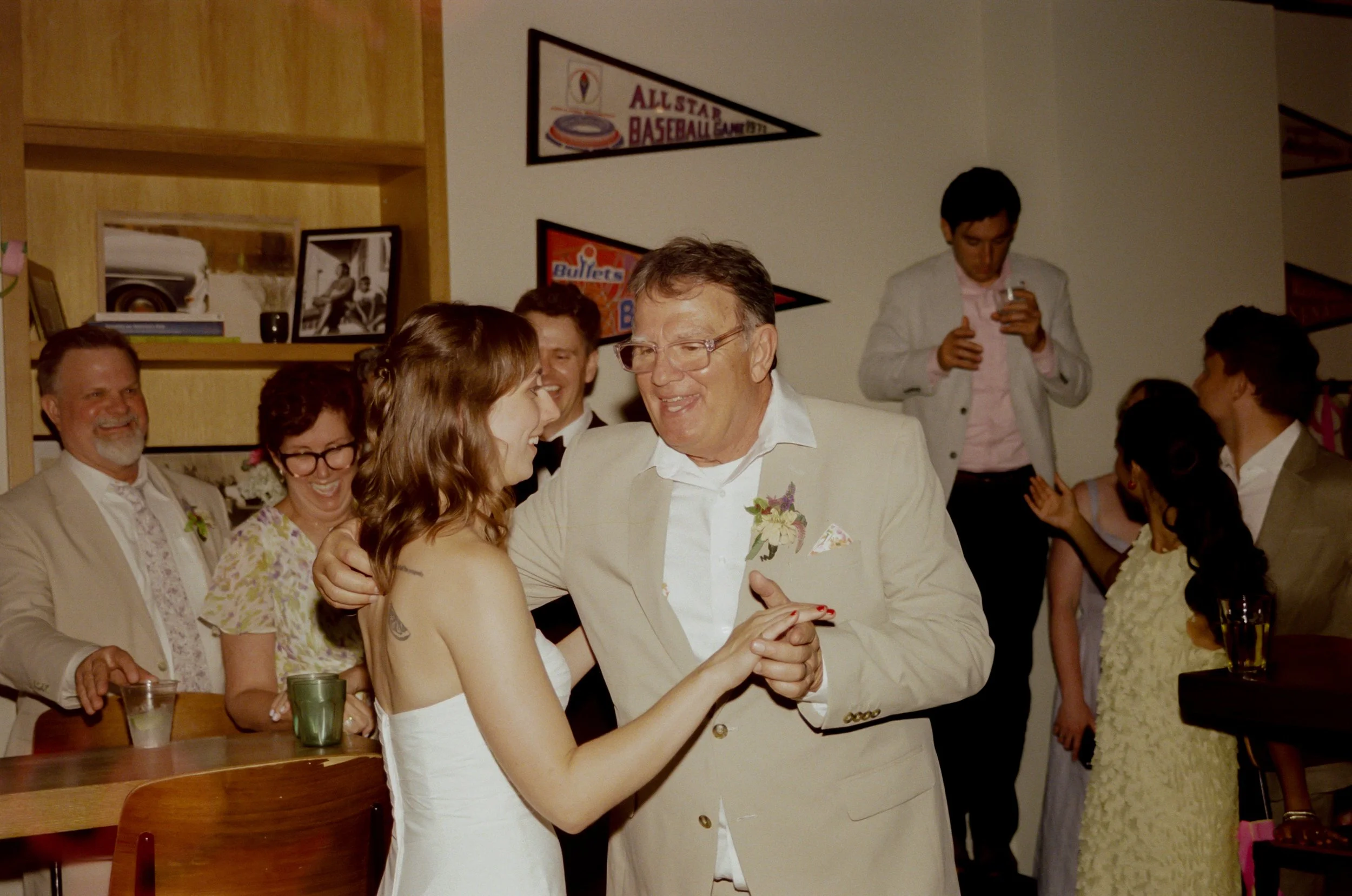 A group of people at a party, including a man in a white suit dancing with a woman in a strapless dress, with others smiling and standing around, some holding drinks.
