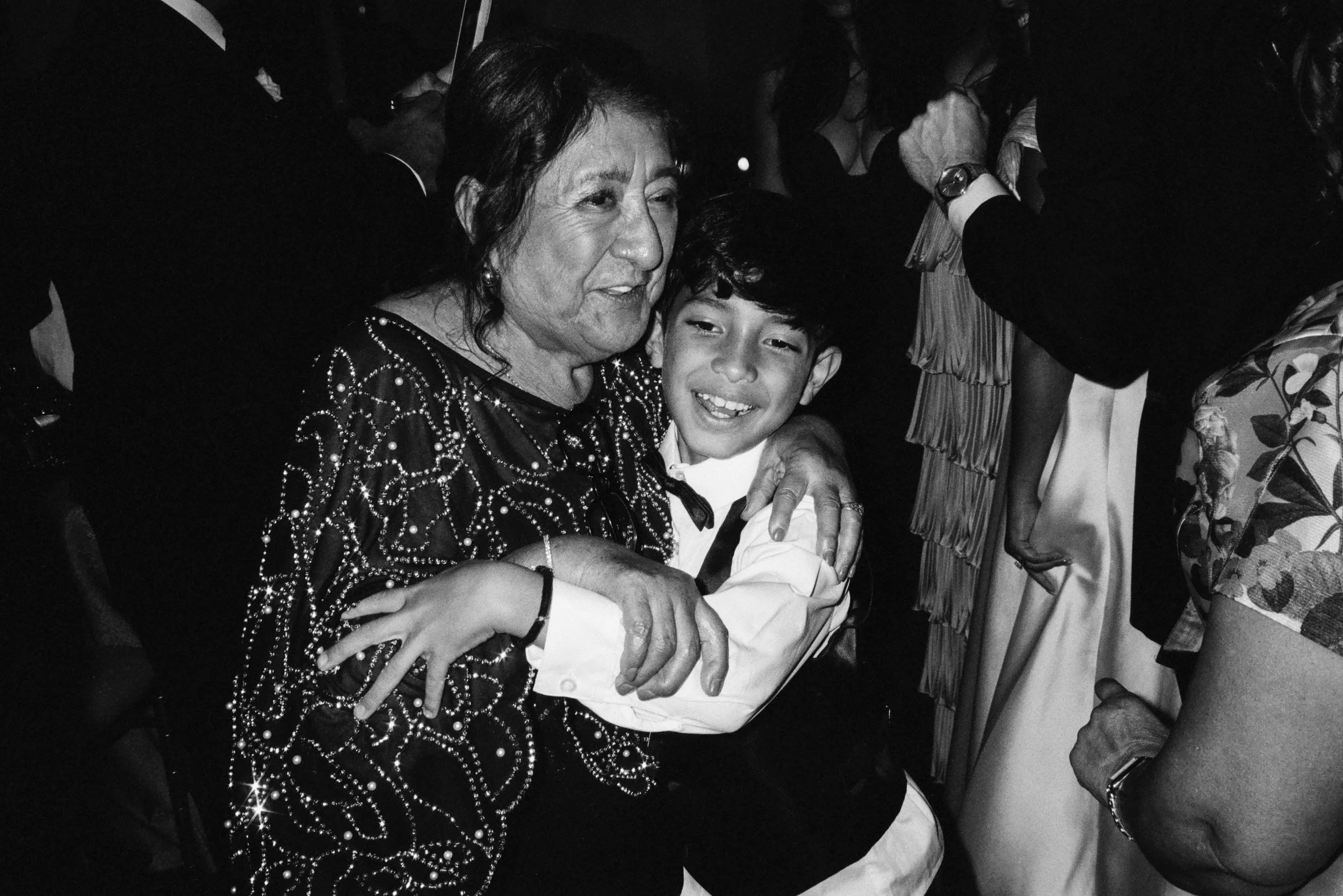 An elderly woman with dark hair styled in a bun, wearing a black dress with a sparkly pattern, is hugging a young boy with short, dark hair, dressed in a white shirt and black bow tie, at a social event. The woman appears to be smiling.