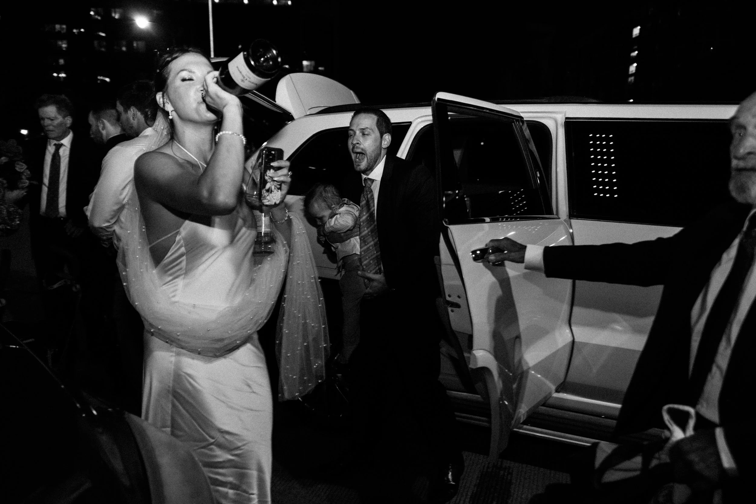 A woman in a wedding dress drinking from a large bottle at night, with several people around her, near an open car door.