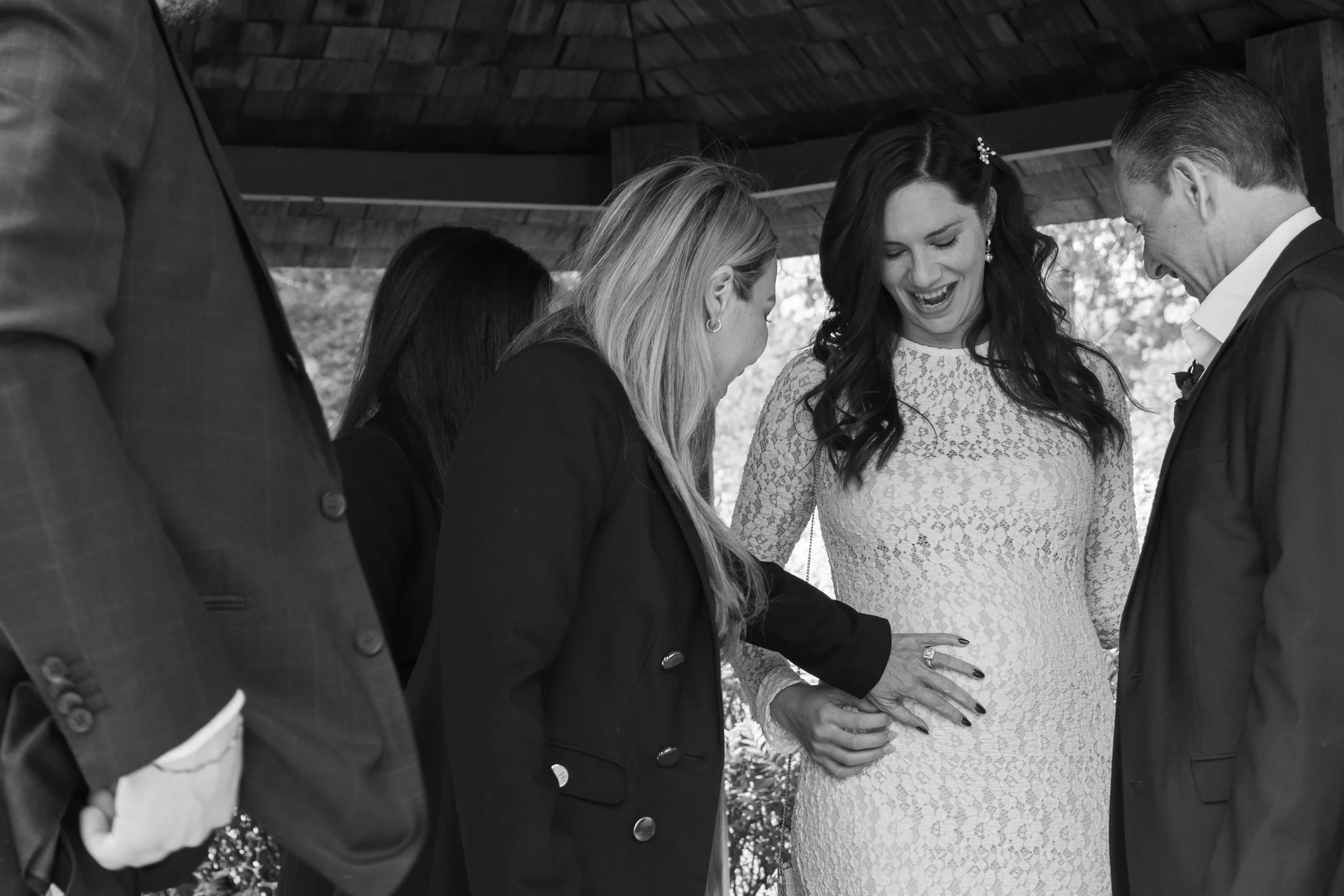 a guest gently touches the brides baby bump while another guest looks on smiling.