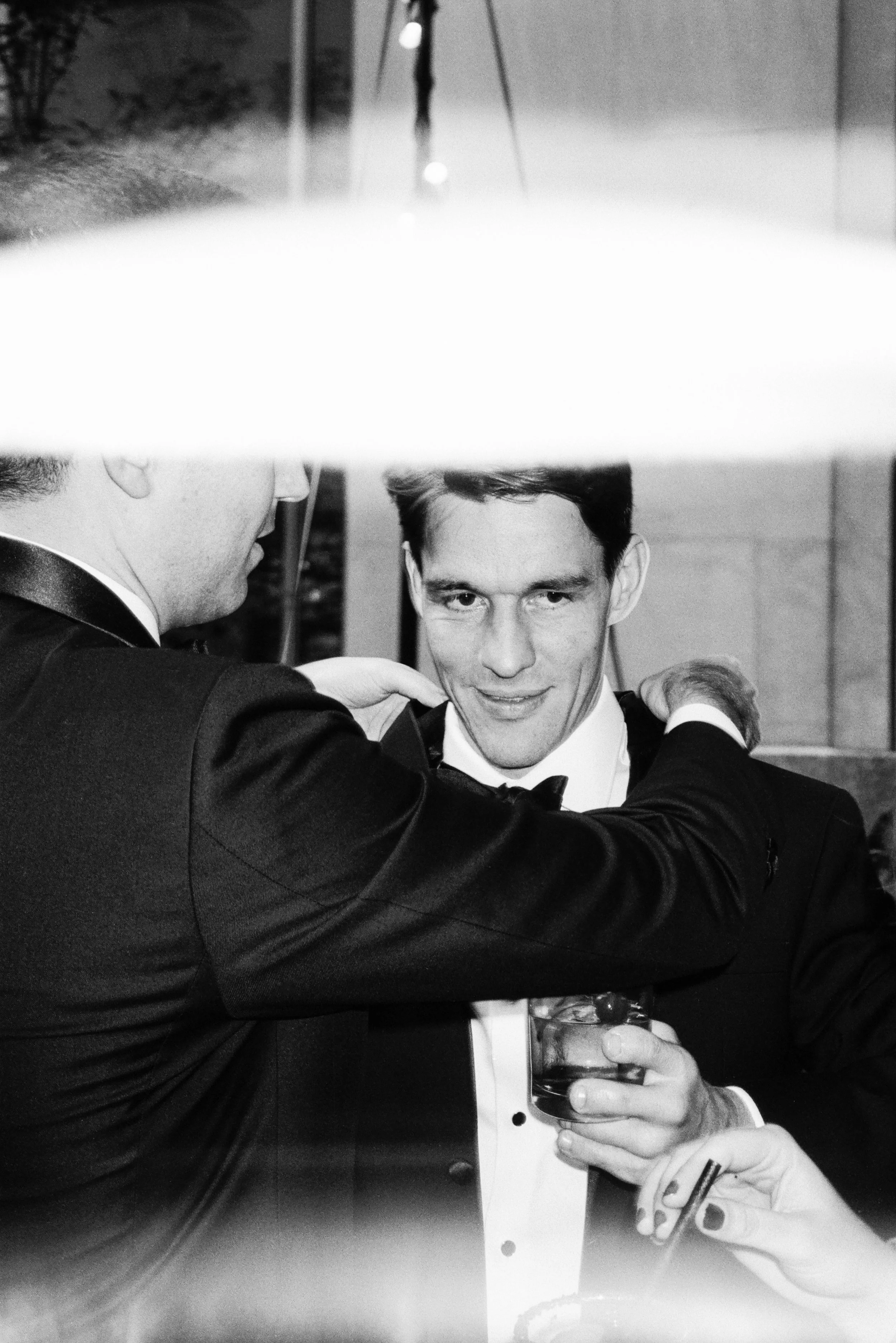 A black-and-white photo of two men, one of whom is taking a selfie with a smartphone. The man in the foreground with dark hair is dressed in a tuxedo and appears to be at a formal event, while the other man has a partially visible face and is also dressed formally. The man in the selfie is holding a glass of beer and smiling.
