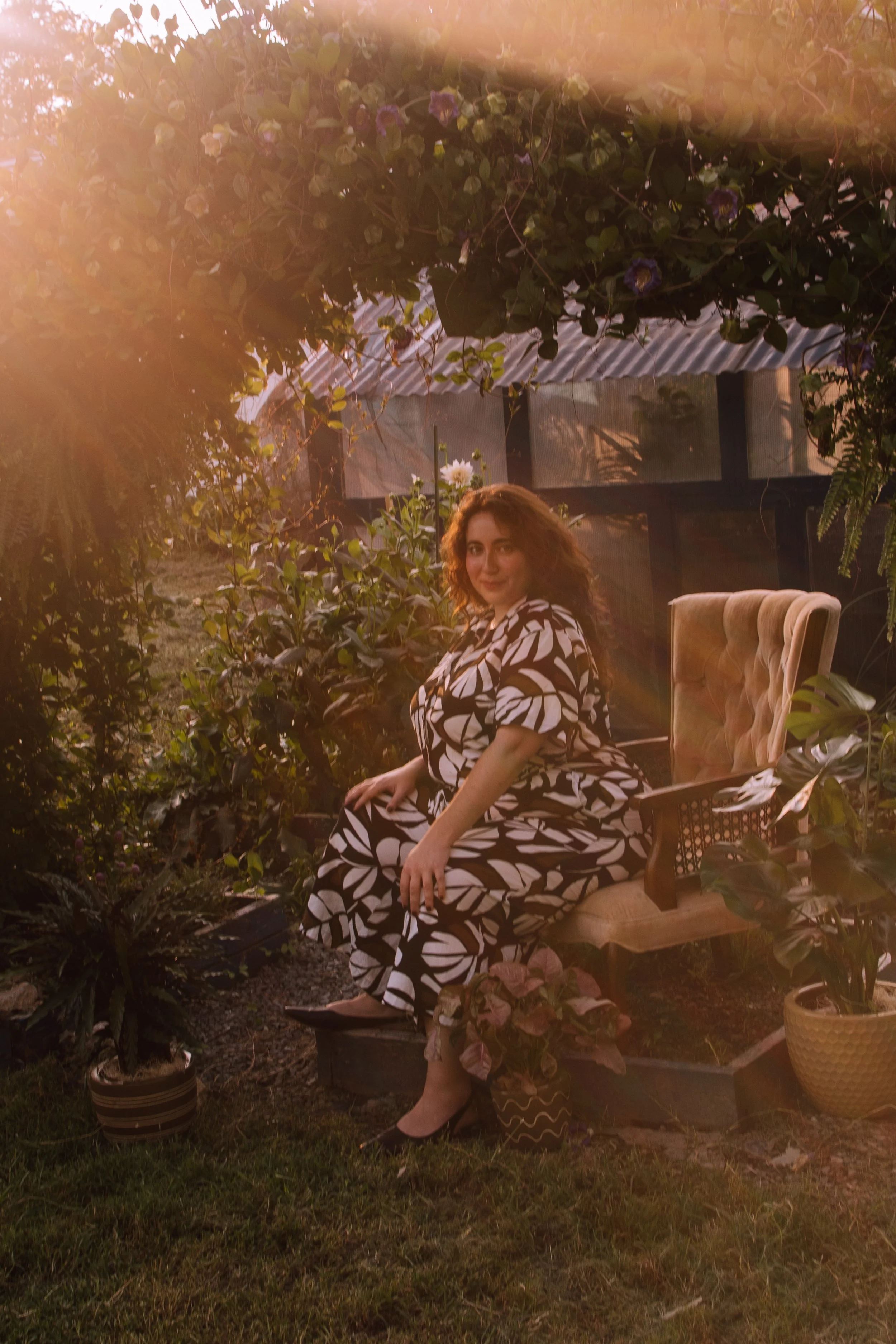 A woman with curly hair wearing a black and white patterned dress sitting on a vintage armchair outdoors, surrounded by plants and flowers during sunset.