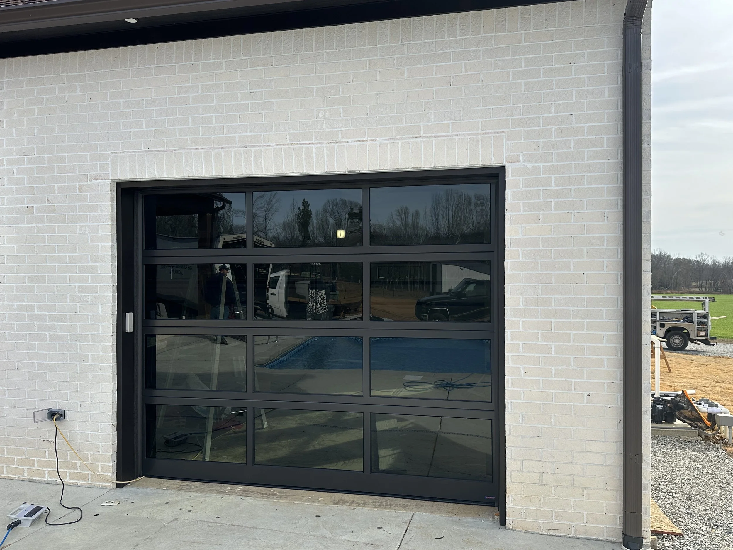 New black garage door with multiple glass panels installed on a white brick wall, with construction equipment and vehicles visible outside.