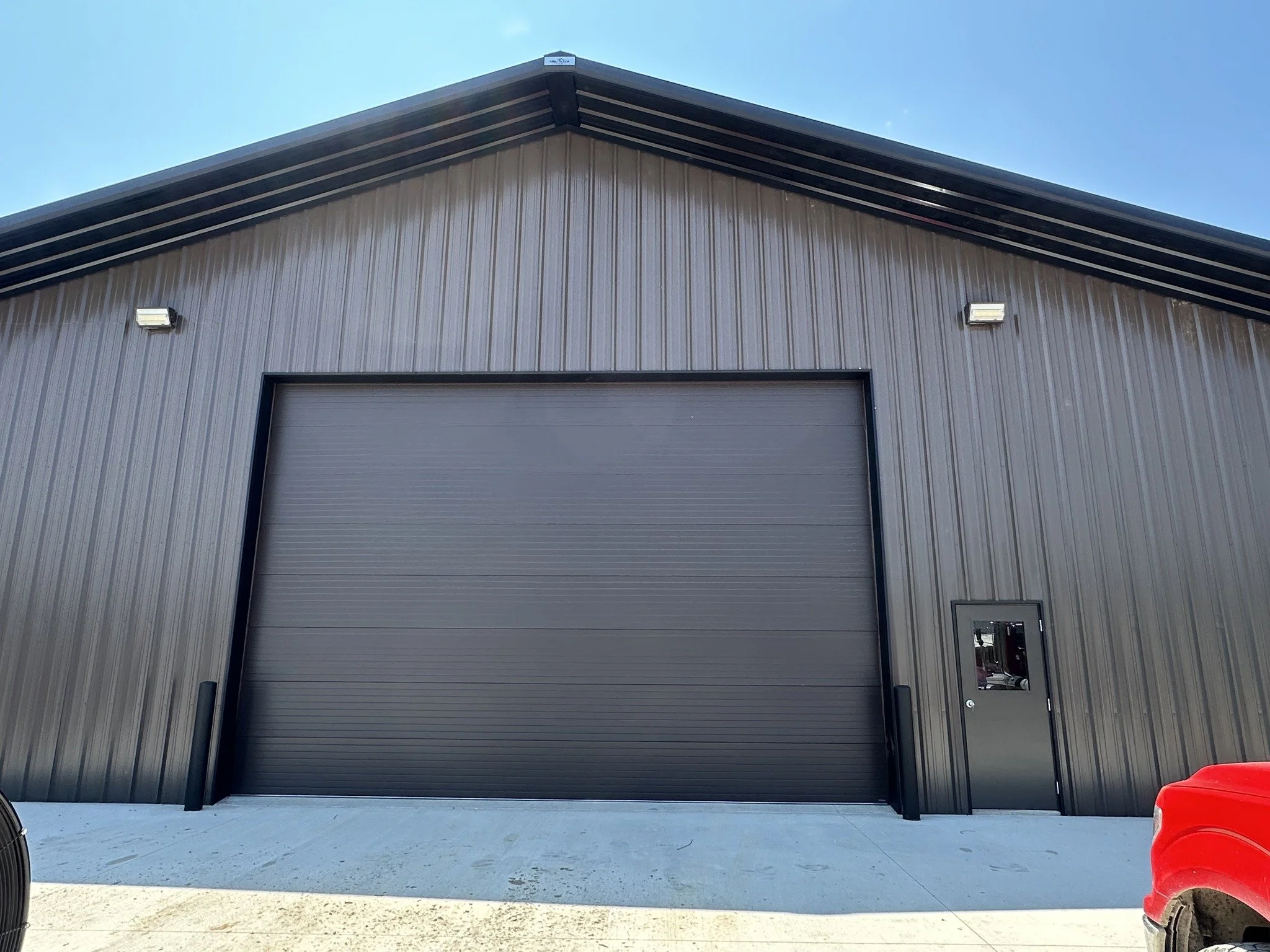 Large industrial building with black metal siding, a large closed gray roll-up door, a small black door with a mirror, and two exterior lamps under a partly cloudy sky.