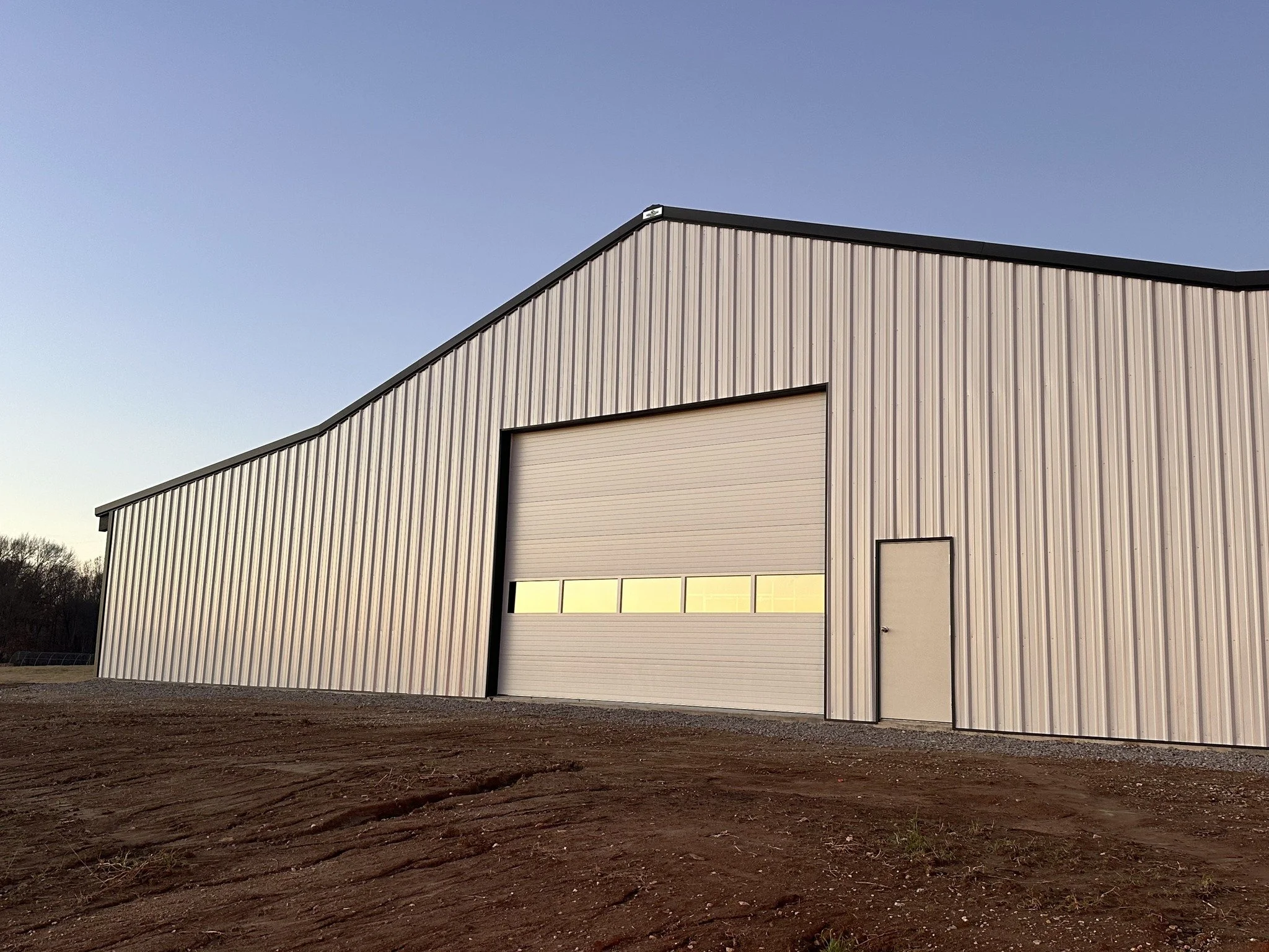 A large metal warehouse building with a closed garage door and a side door, set on dirt ground, under a clear sky.
