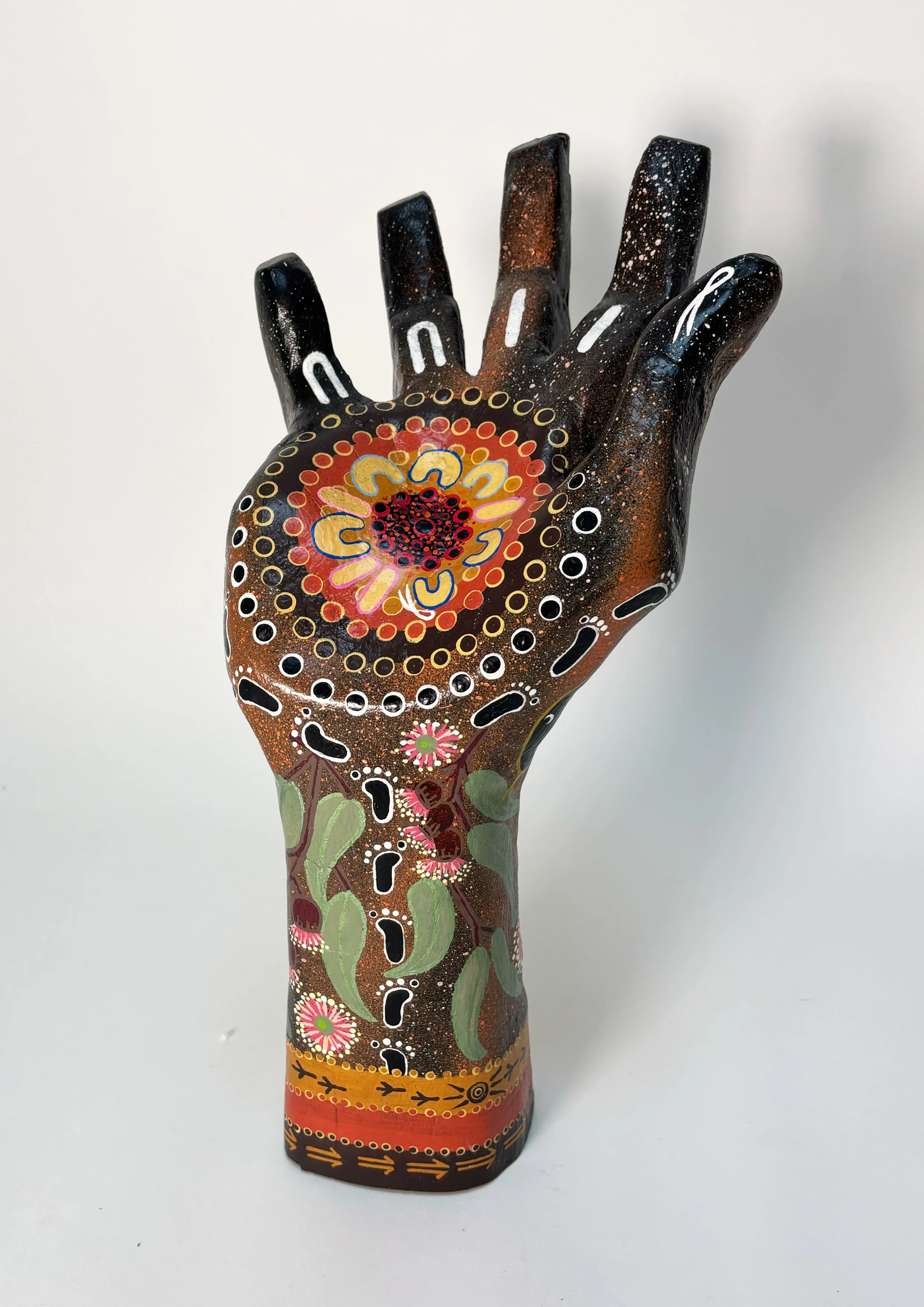 A hand-shaped sculpture painted with intricate, colorful patterns including flowers, dots, and tribal designs on a white background. Indigenous Art, community driven