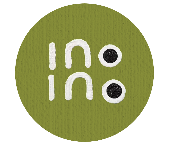 Close-up of a green circular button with the phrase 'in no' written twice, with each 'in' paired with a black dot, creating a stylized design. Aboriginal community art