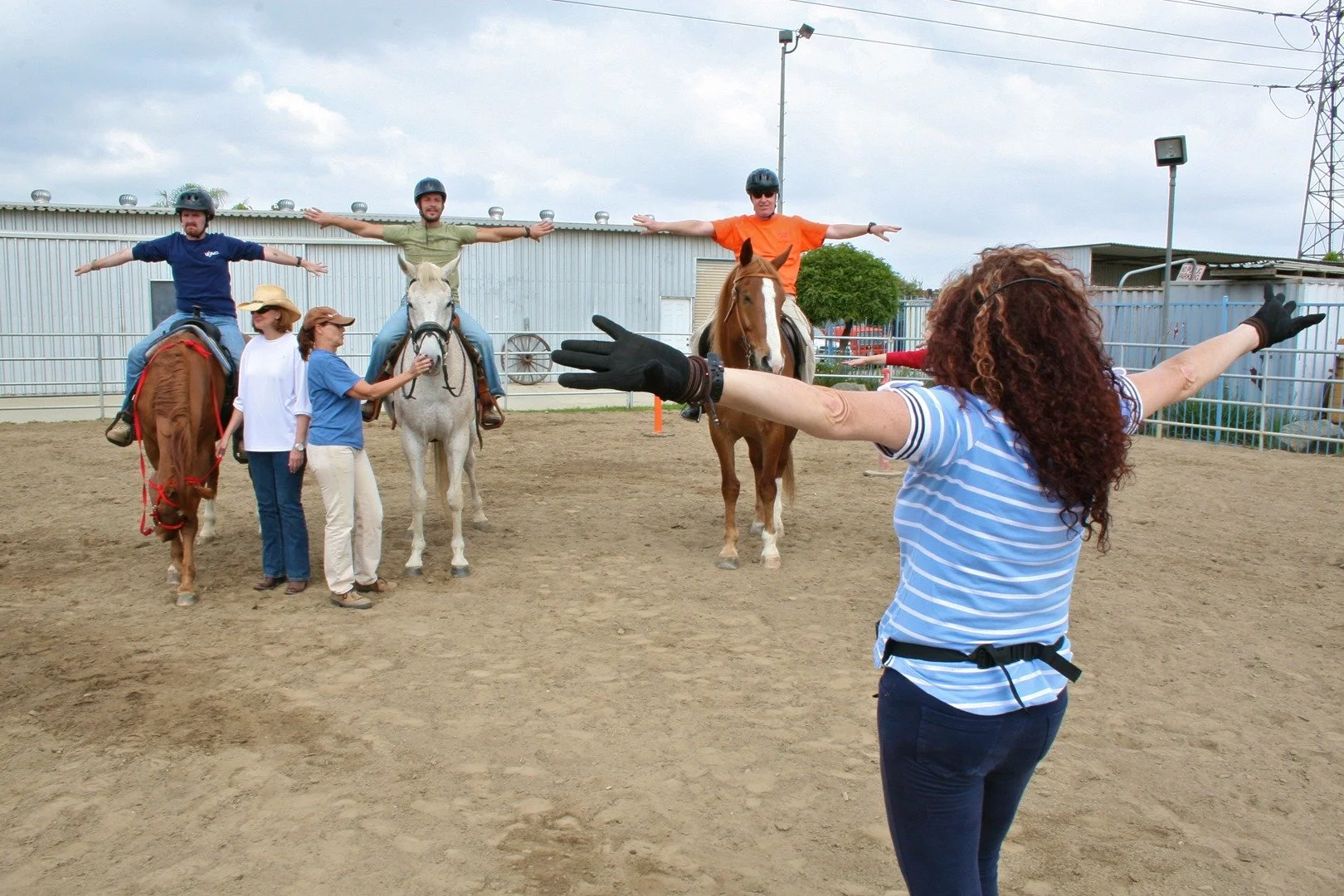 Three people on horseback with arms outstretched, horses standing on a dirt area, woman with curly red hair standing in front with arms outstretched, in an outdoor equestrian facility.