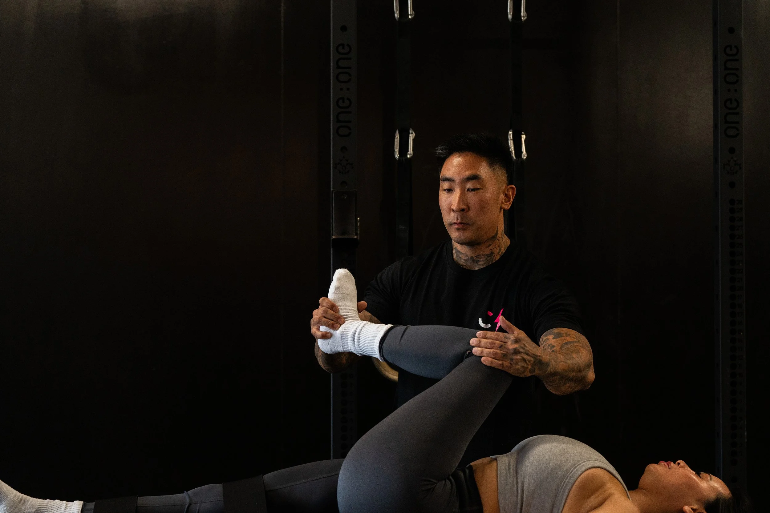 A man assisting a woman with a leg exercise in a gym, with black workout equipment in the background.