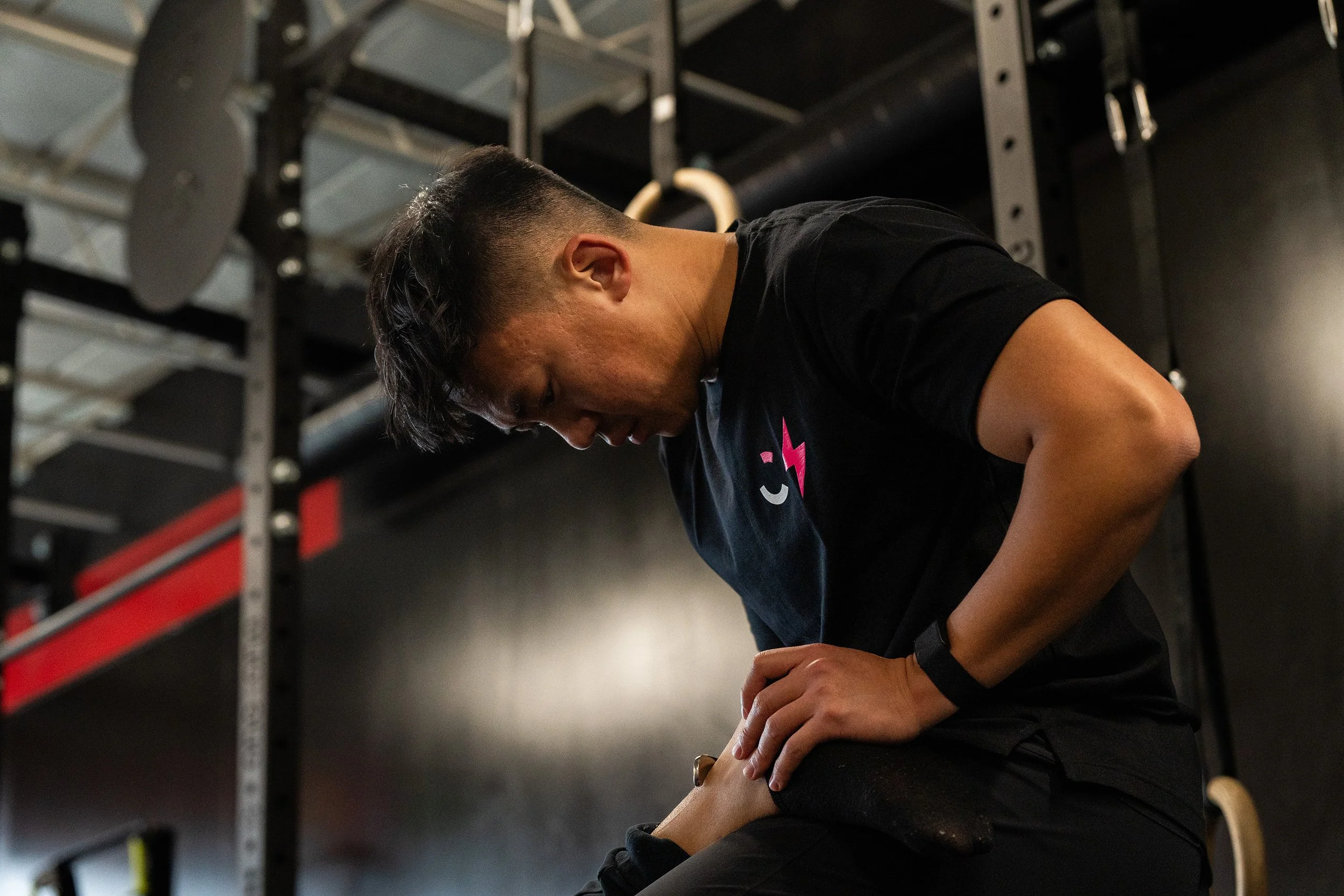 A man in a gym, sitting on a stool, stretching his ankle, wearing a black t-shirt with a pink graphic, with gym equipment in the background.