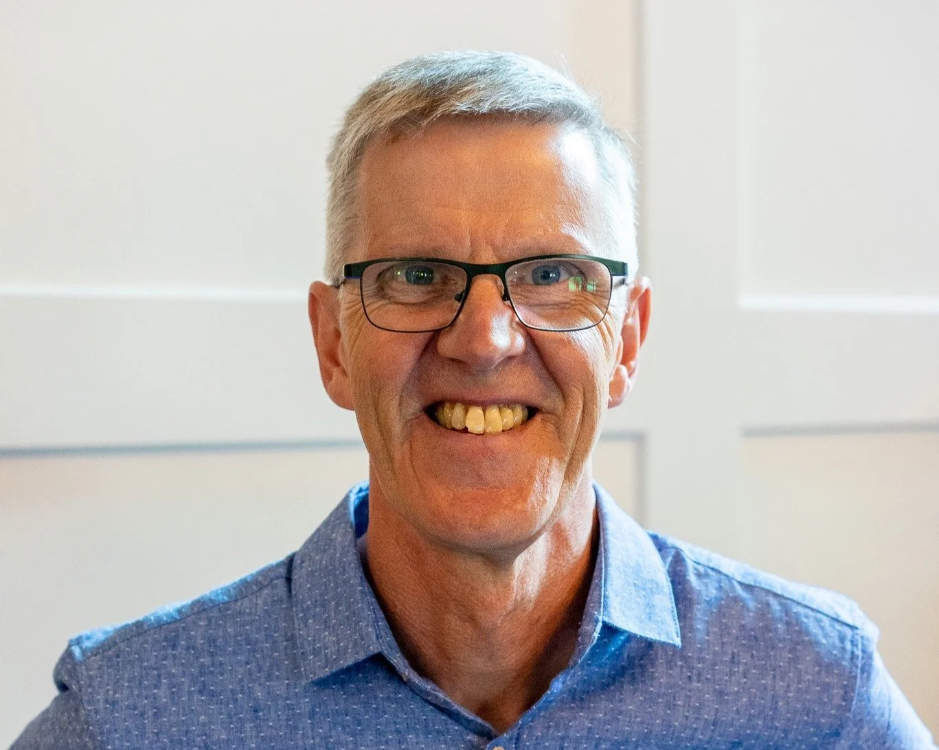 Smiling man with glasses, short gray hair, wearing a blue button-up shirt, indoors with white wall background.