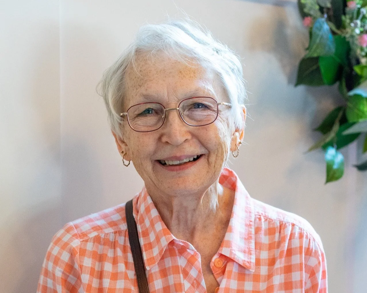 A smiling elderly woman with white hair, wearing glasses, earrings, and a pink checkered shirt, standing indoors with green foliage and flowers in the background.
