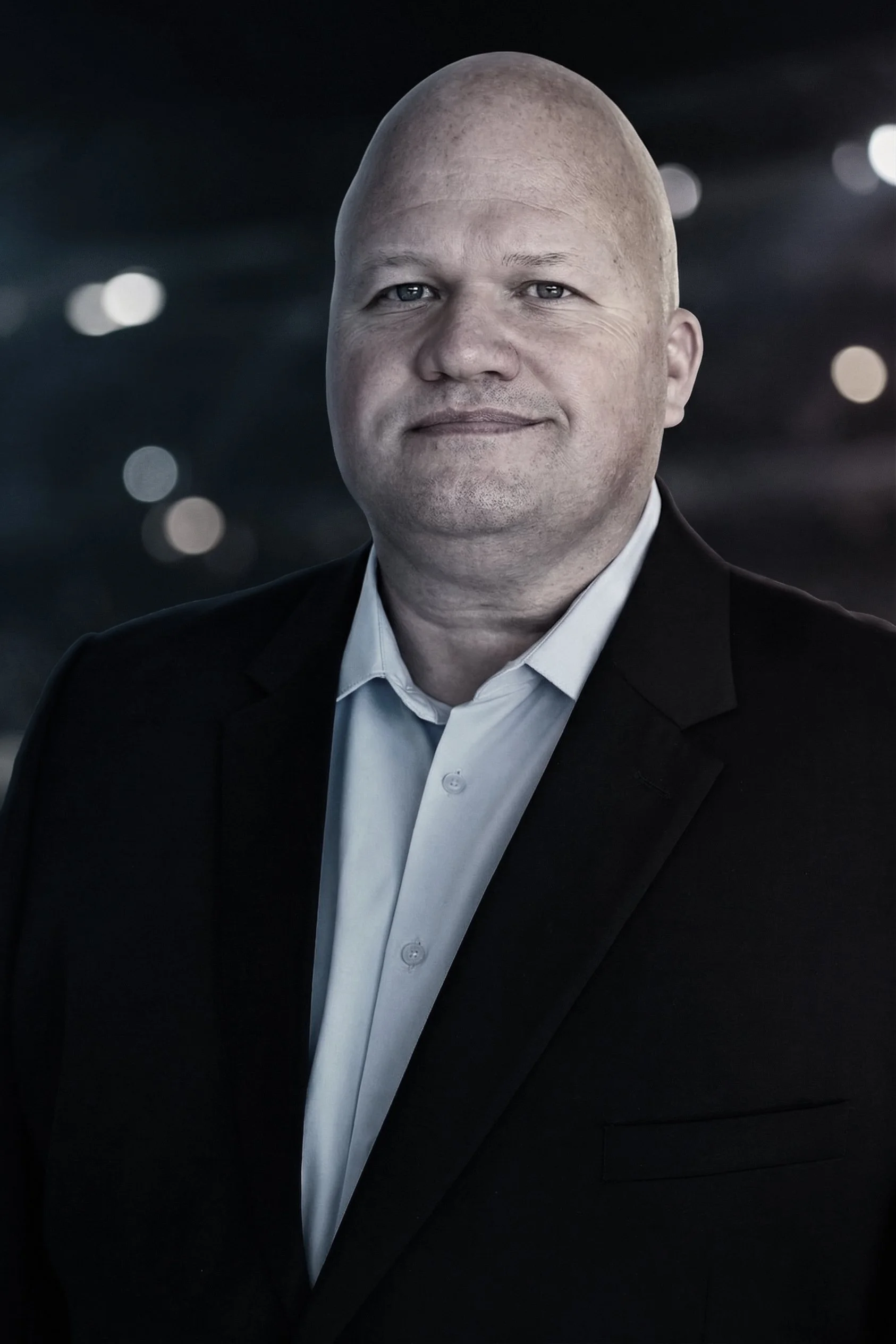 A bald man in a white shirt and black suit jacket standing against a dark blurred background with circular light spots.