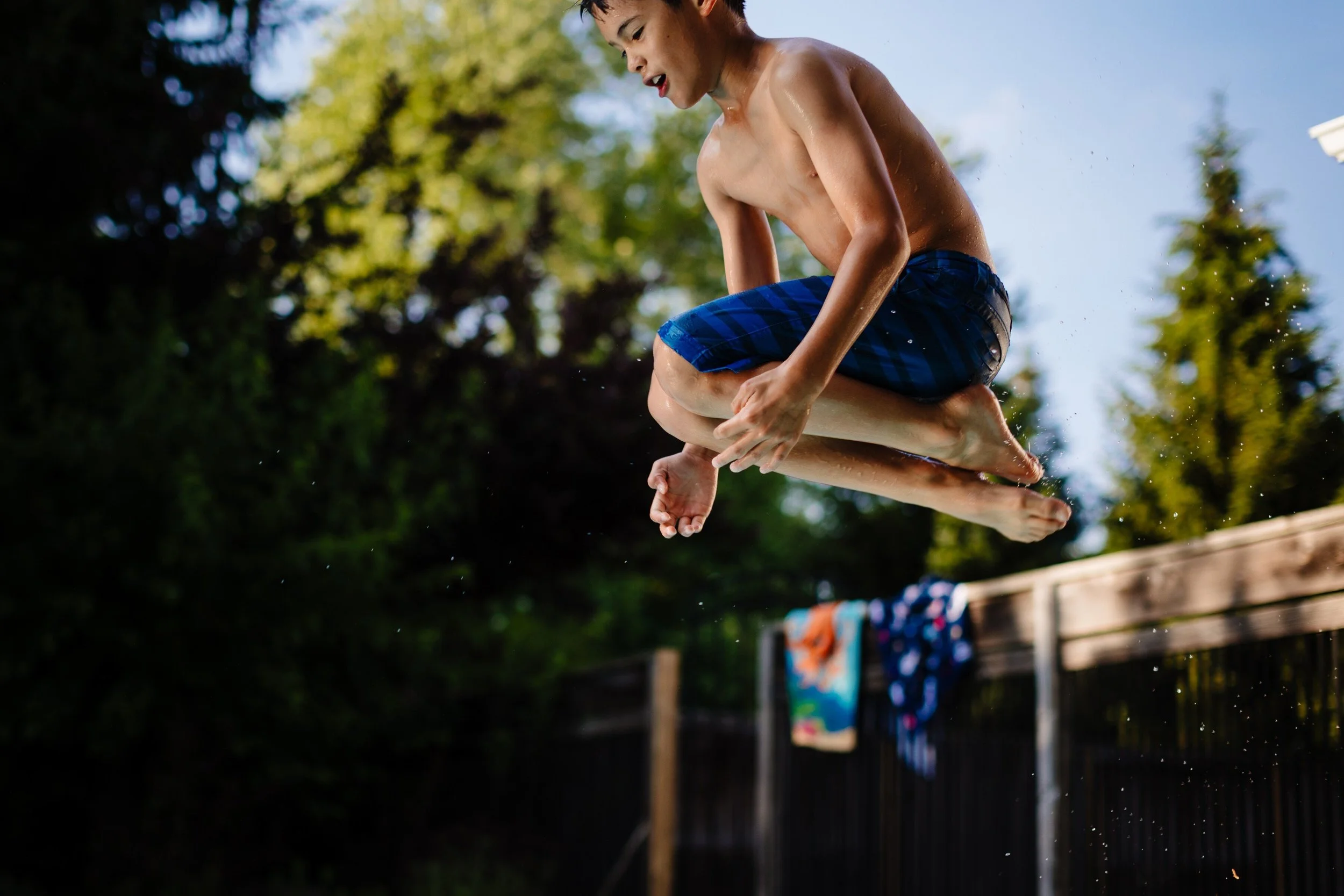 A young boy jumping into a swimming pool outdoors with sunlight shining and trees in the background.