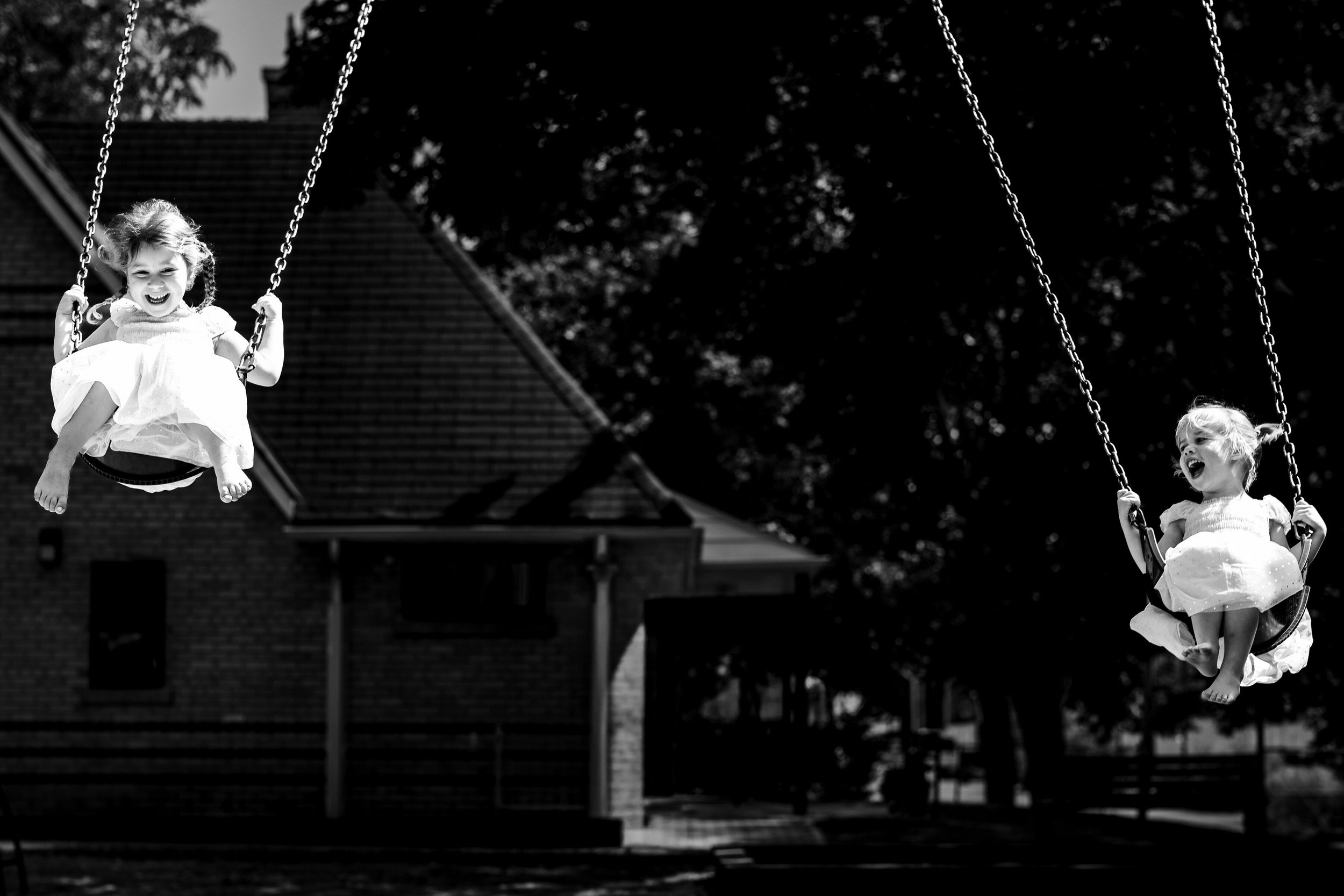 Two young girls are playing on swings in a playground, with trees and a house in the background. Both girls are smiling and having fun.