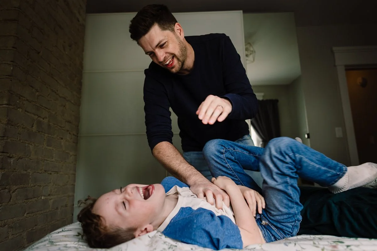 A man playfully wrestling with a young boy on a bed in a bedroom with a brick wall.