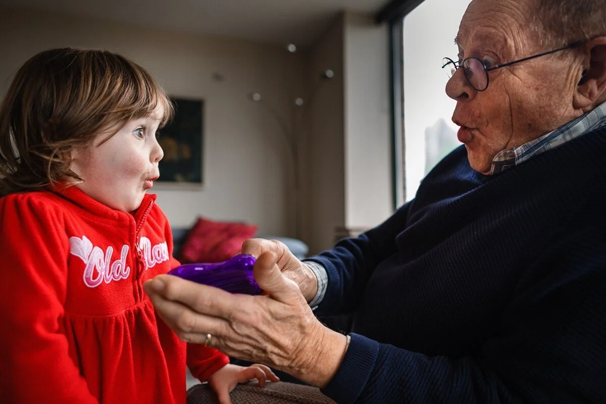 An elderly man and young girl sitting together indoors, both looking surprised.