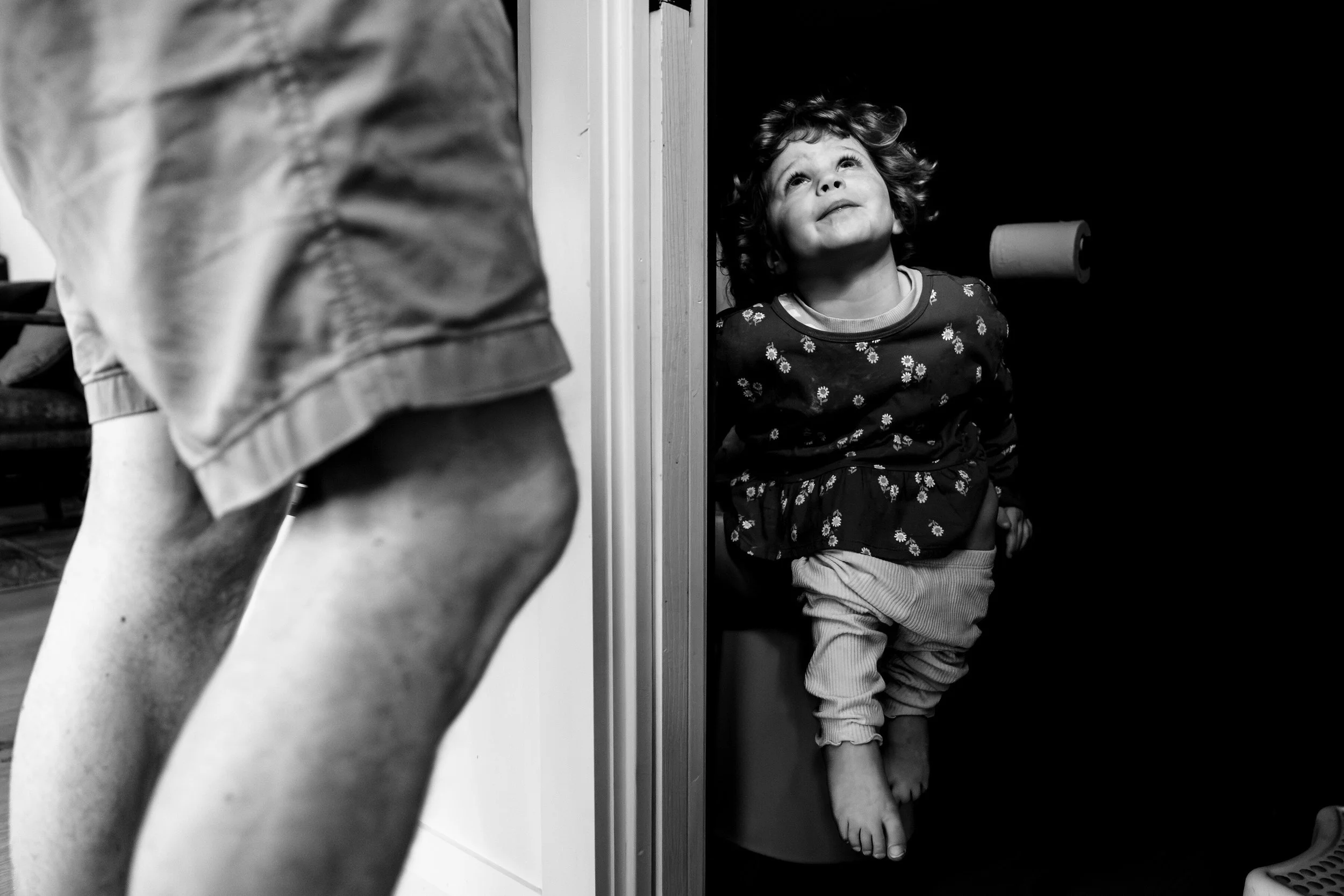 A young girl with curly hair looking up and smiling while sitting on a toilet in a dark bathroom. An adult's legs and a blurry lower body are partially visible in the foreground.