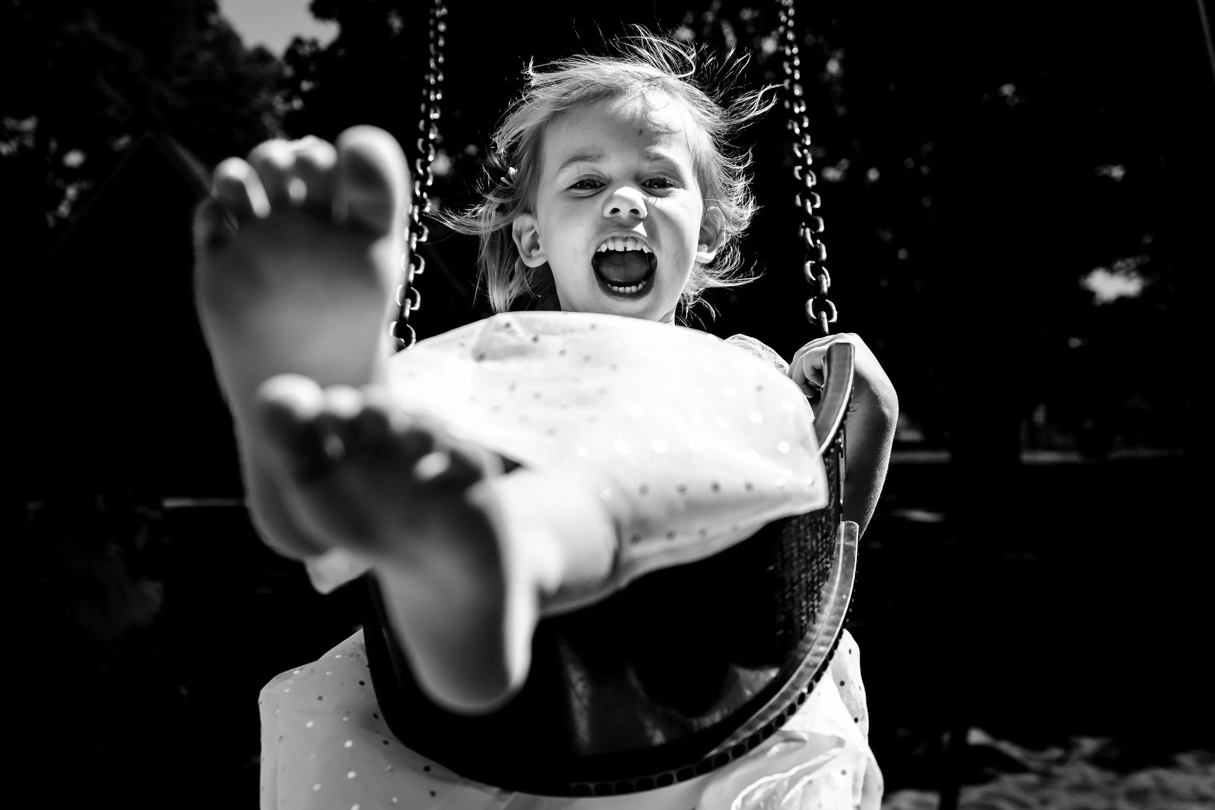 Black and white photo of a young child on a swing, looking directly at the camera with an open mouth, outdoors with trees in the background.
