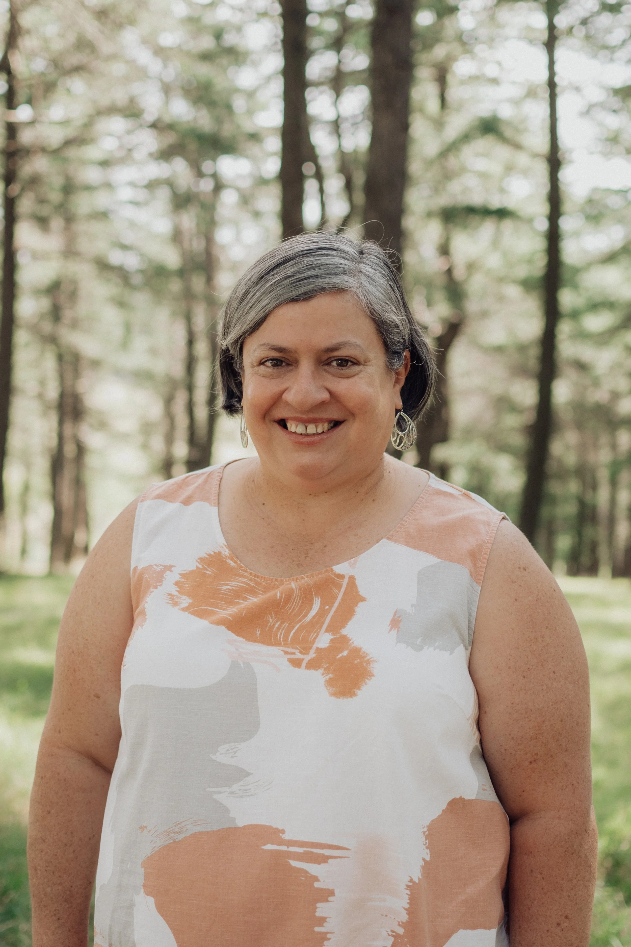 A woman with short gray hair smiling outdoors in a forested area, wearing a sleeveless dress with an abstract orange, gray, and white pattern.
