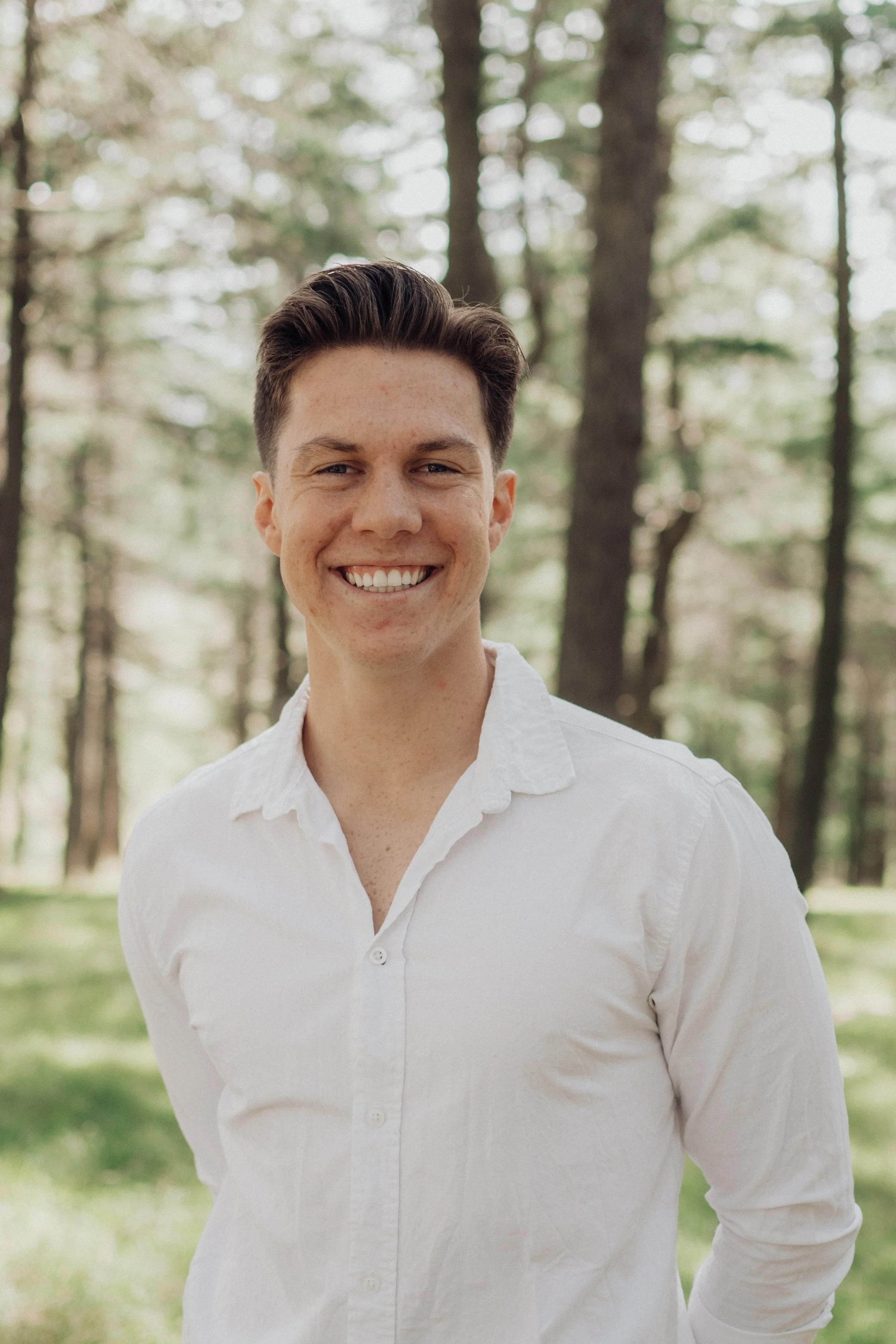 A smiling young man with short dark hair wearing a white button-up shirt standing outdoors in a wooded area.