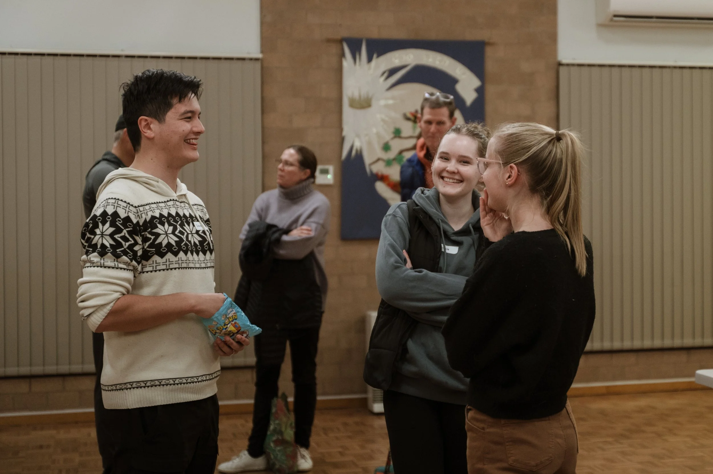 Group of young people smiling and chatting indoors, with a couple women engaged in conversation and a young man holding a snack bag, while a woman in the background looks on.