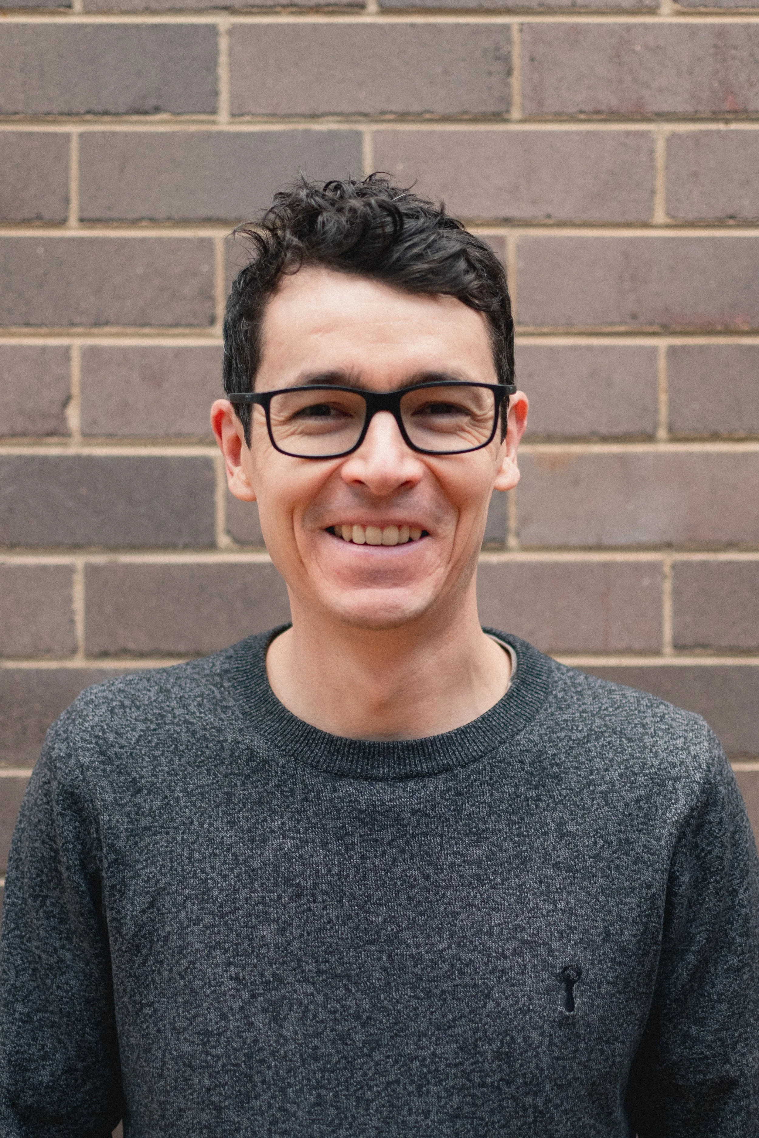A smiling man with short dark hair, glasses, and a dark gray sweater, standing in front of a brick wall.