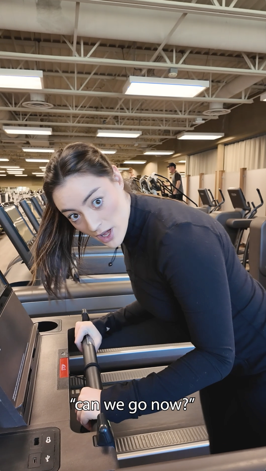 A young woman with brown hair looking surprised while holding onto a treadmill at a gym, with exercise equipment and other people in the background.