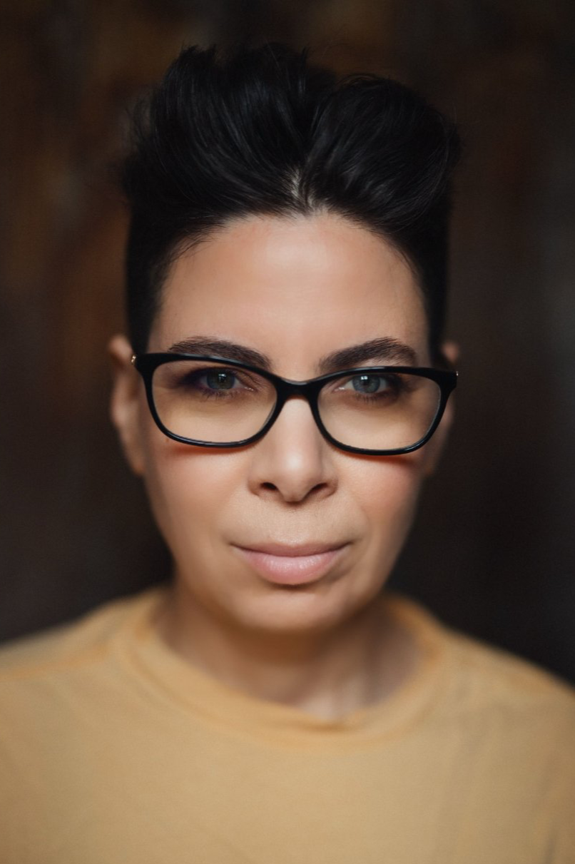 Close-up portrait of a woman with short dark hair, wearing black glasses and a beige top, looking directly at the camera with a neutral expression.