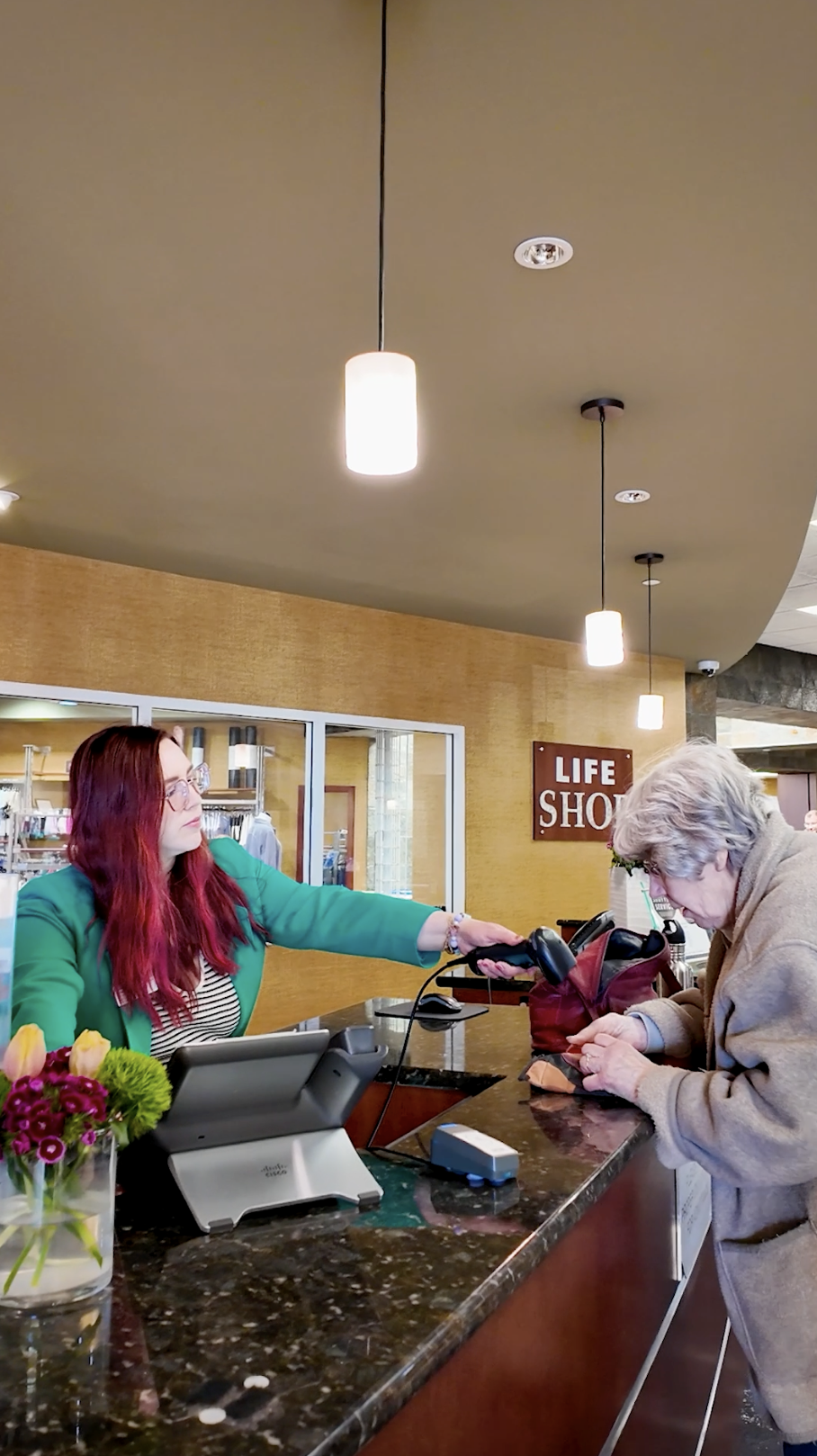 A woman at a hotel reception desk handing a card or key to an older woman who is checking in. The reception area has a granite counter, a vase with flowers, and a sign that reads 'LIFE SHOT'. The ceiling has hanging lights.