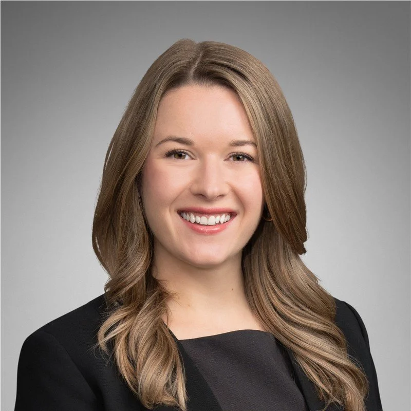 A smiling woman with shoulder-length light brown hair, wearing a black blazer, against a neutral gray background.