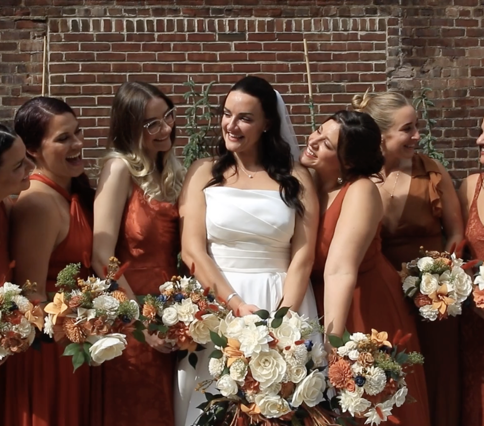 Bride in white surrounded by bridesmaids in red dresses holding floral bouquets against a brick wall.