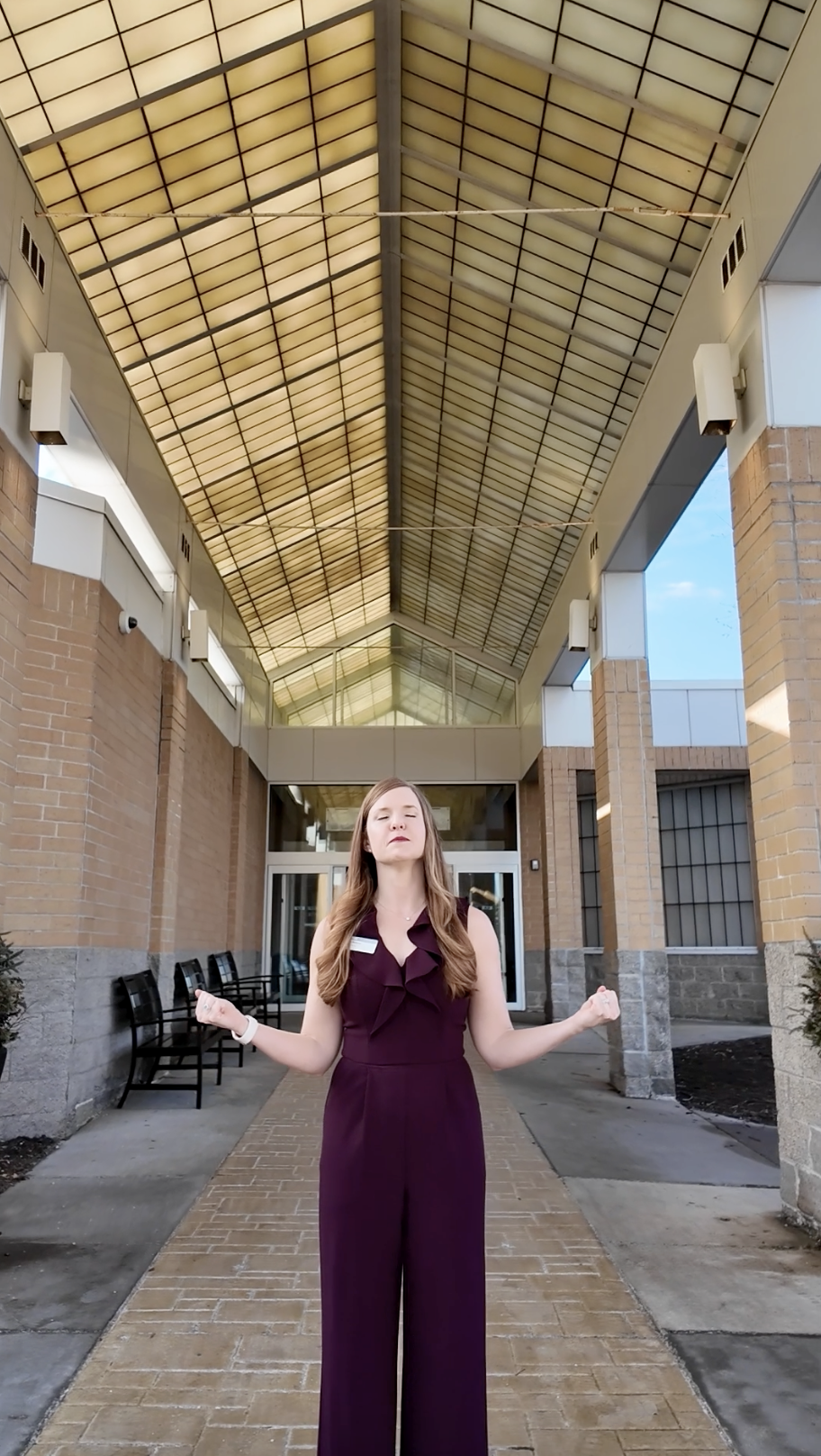 Woman standing outdoors under a large, covered walkway with a glass and metal ceiling. She is wearing a sleeveless, dark purple jumpsuit, has her eyes closed, and appears to be meditating with her arms raised and fists clenched.