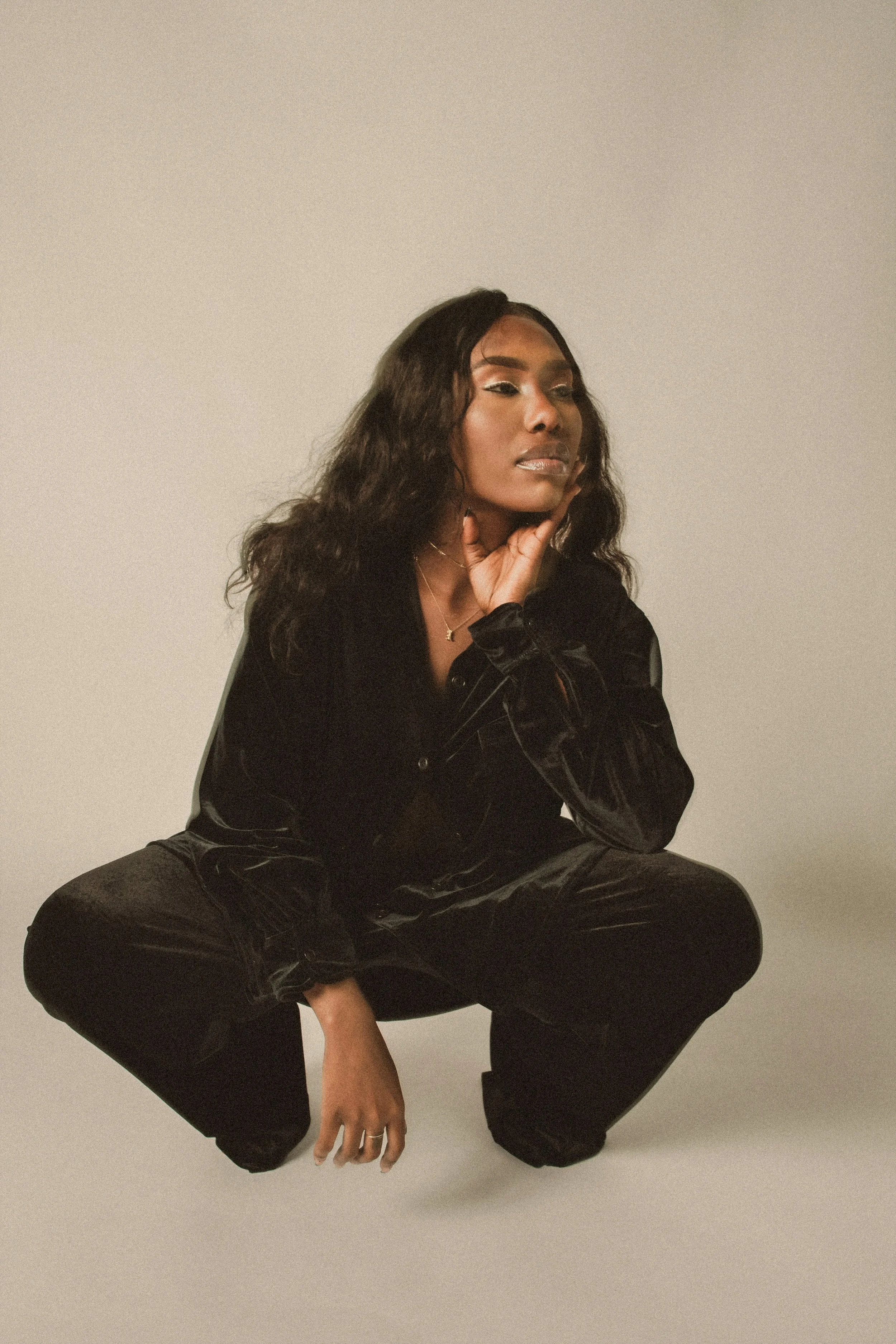 A woman in black clothing is squatting on the floor, looking to her left with her hand touching her neck. The background is plain and off-white.