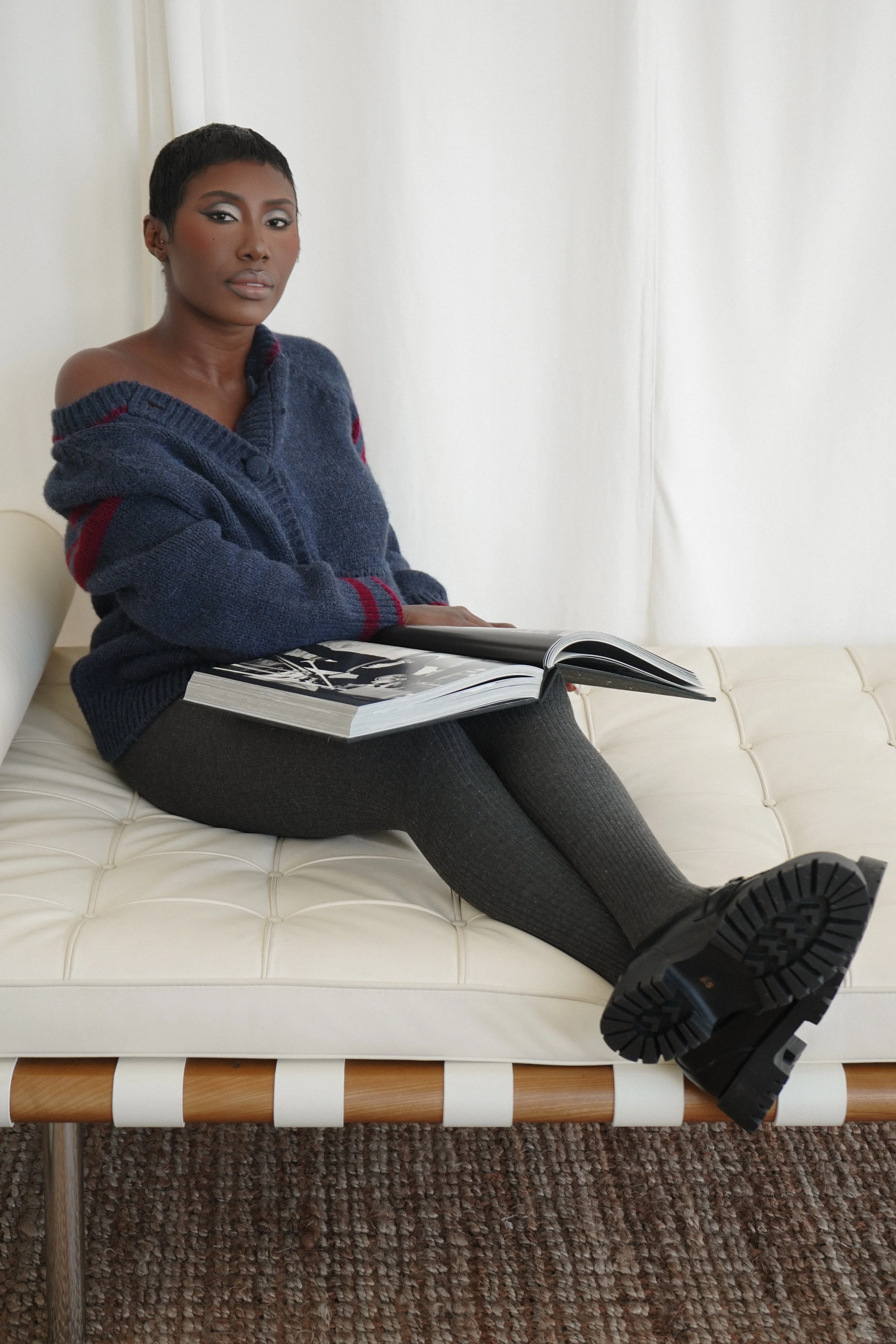 A woman with short black hair and dark skin wearing a blue sweater, black leggings, and black boots sitting on a white tufted bench, holding an open magazine, in a room with white walls and a textured brown carpet.
