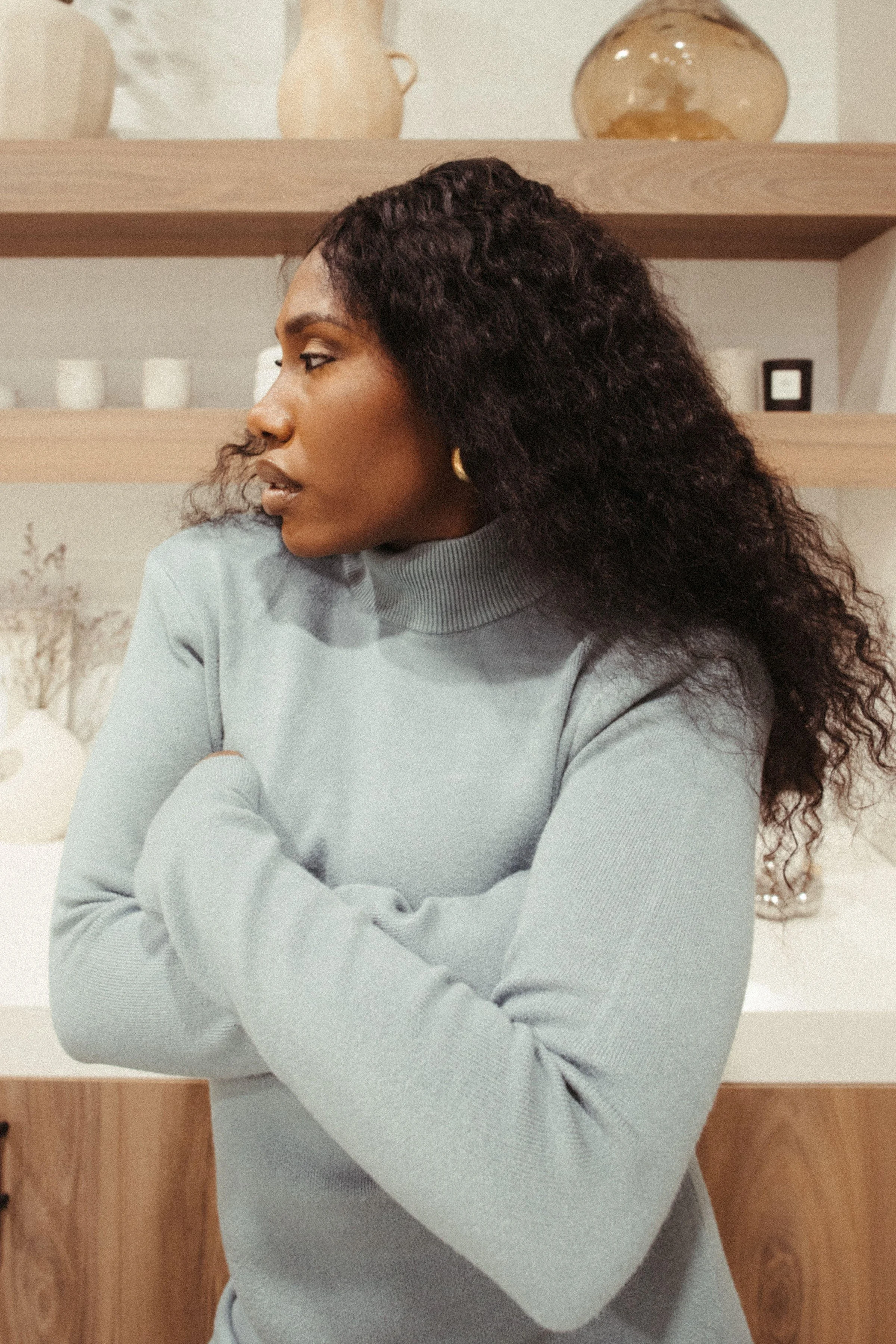 A woman with curly hair wearing a light gray turtleneck sweater standing indoors with wooden shelves and decorative items in the background.