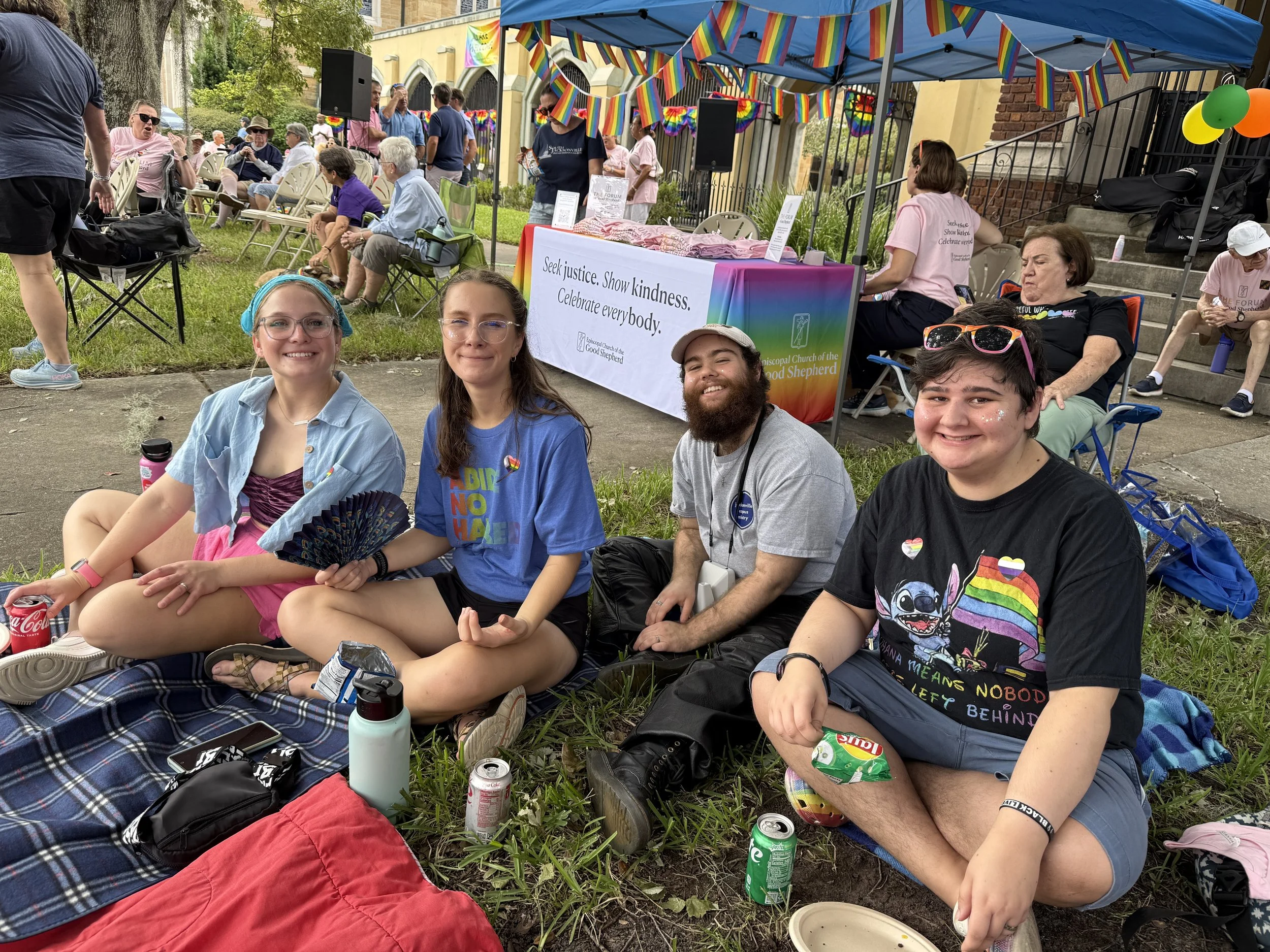 Four smiling individuals sitting on grass at a pride event, with rainbow decorations and a table in the background with a rainbow banner. They are surrounded by drinks and personal items.