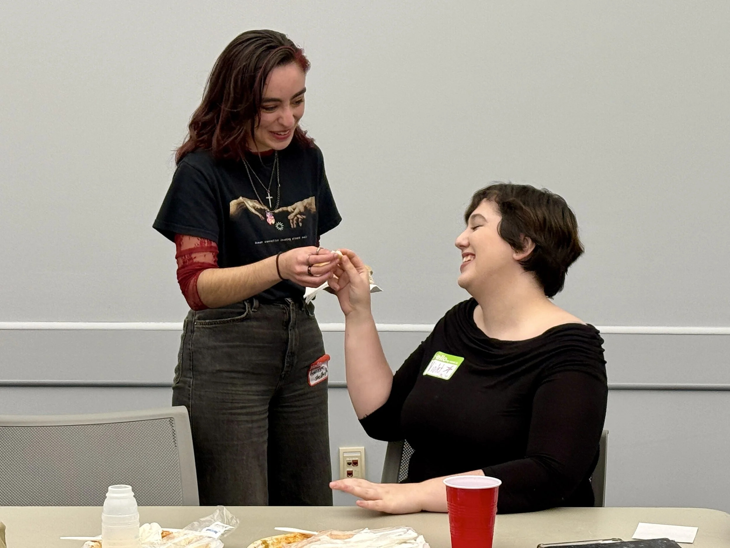 A person with brown hair and a black t-shirt handing a small object to a person with short dark hair sitting at a table, both smiling.