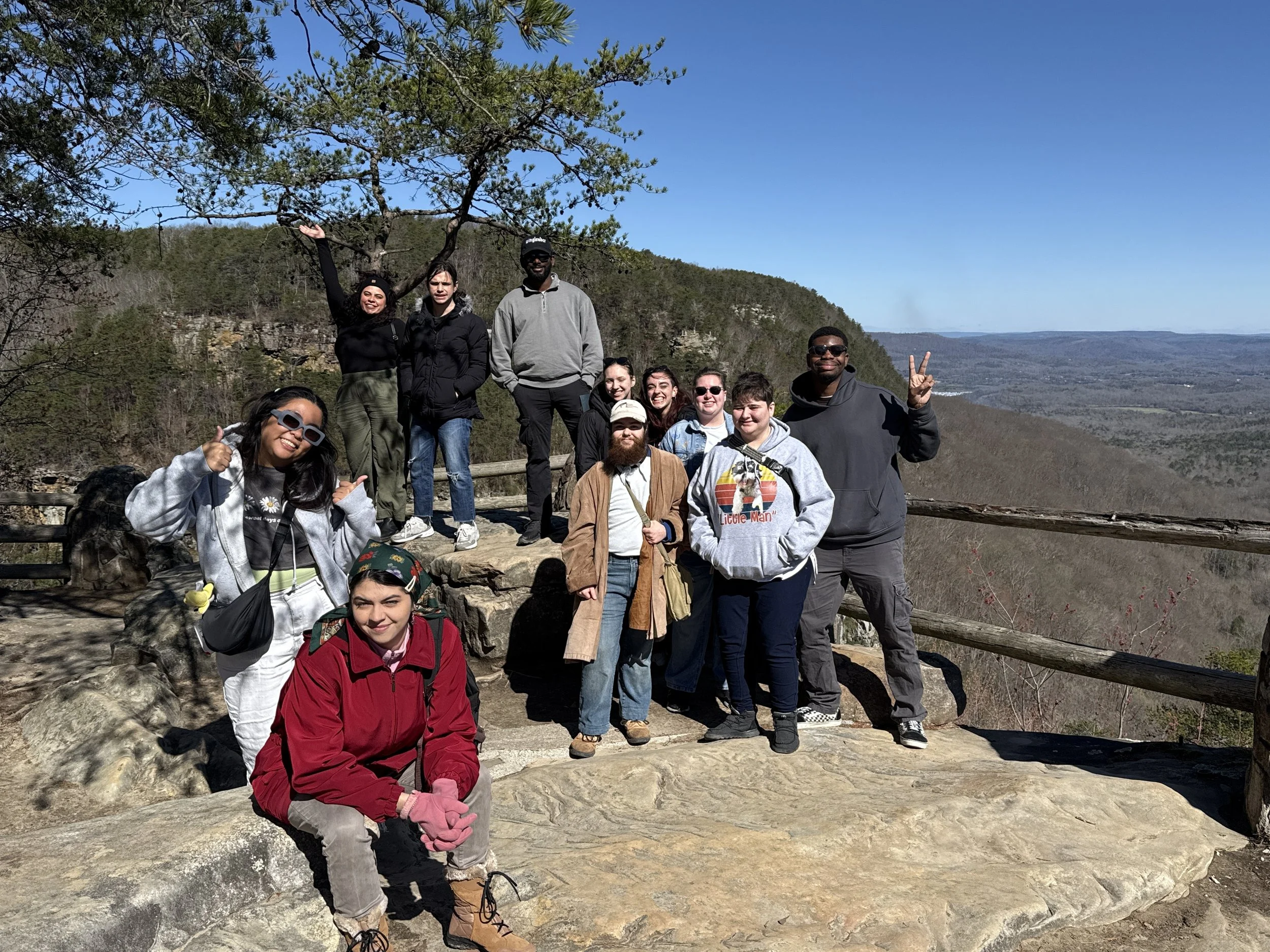 A group of fifteen people posing on a rocky overlook in a forested area with a scenic view of distant hills and valleys on a sunny day.